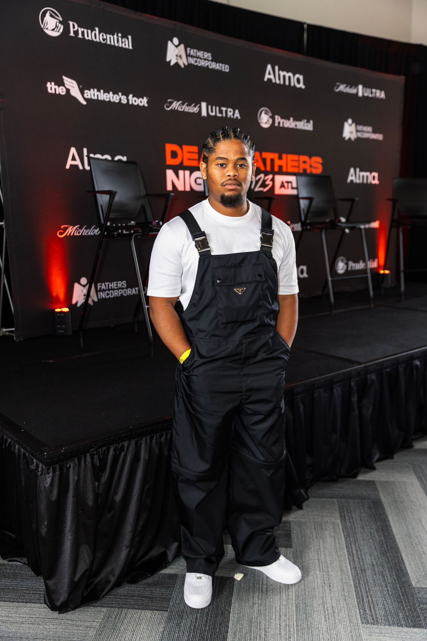 A person wearing overalls and white t-shirt poses at a branded event backdrop.