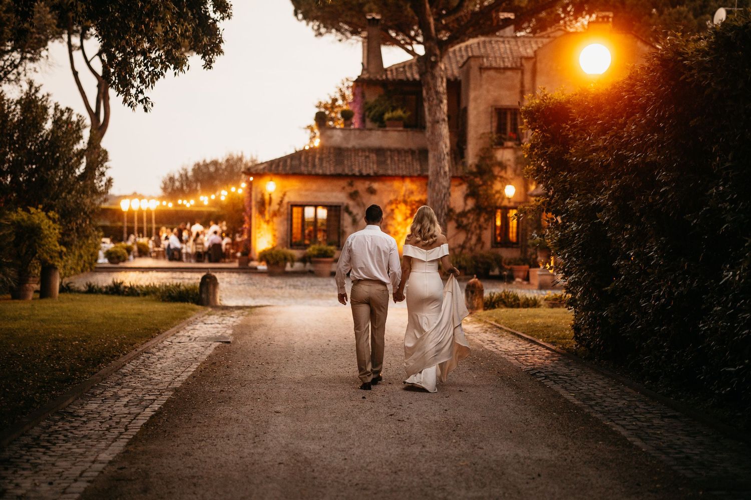 A couple walks hand in hand down an evening pathway with warm street lights glowing at sunset.