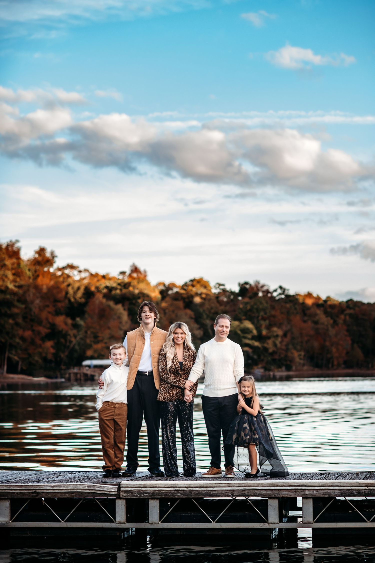Family of five poses on a wooden dock at sunset with autumn trees and a lake in the background.