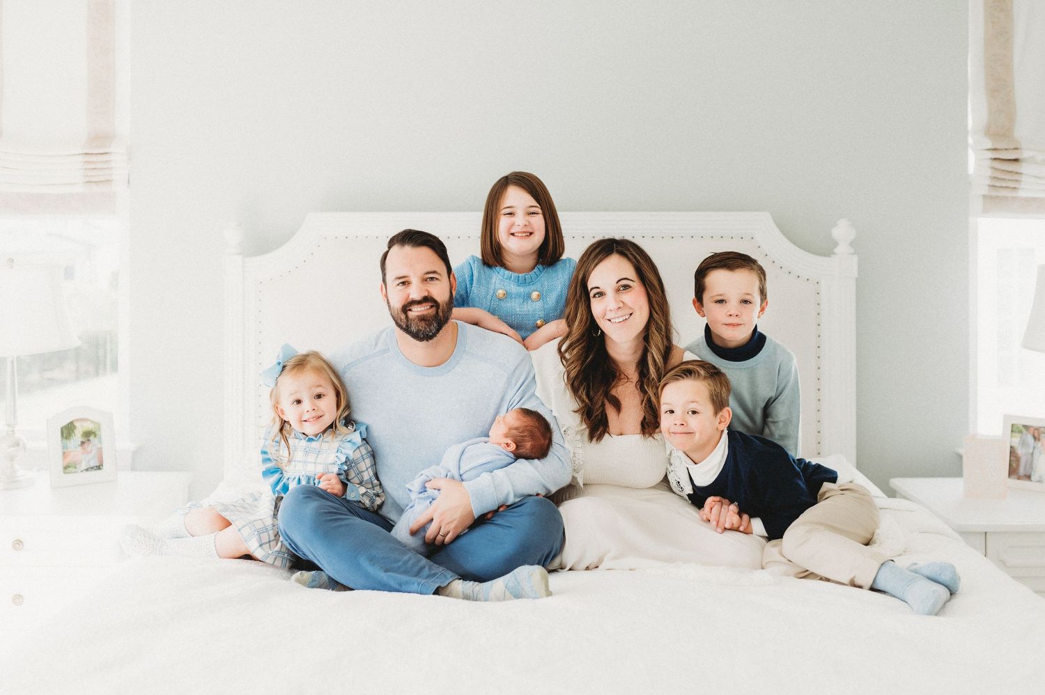 A family of seven sits together on a white bed wearing coordinated casual outfits in a bright bedroom setting.