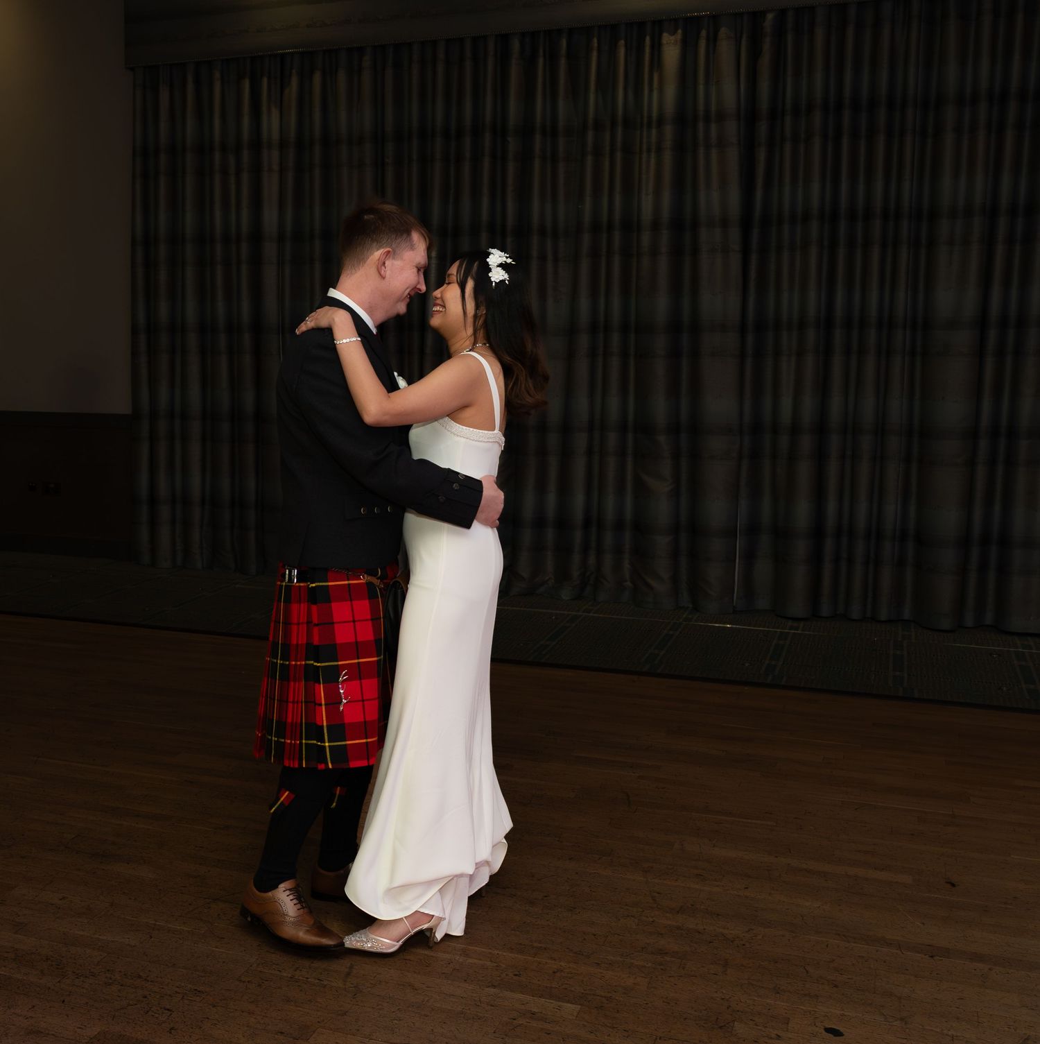 Two dancers performing a romantic dance in white dress and plaid kilt on a dark wooden dance floor.