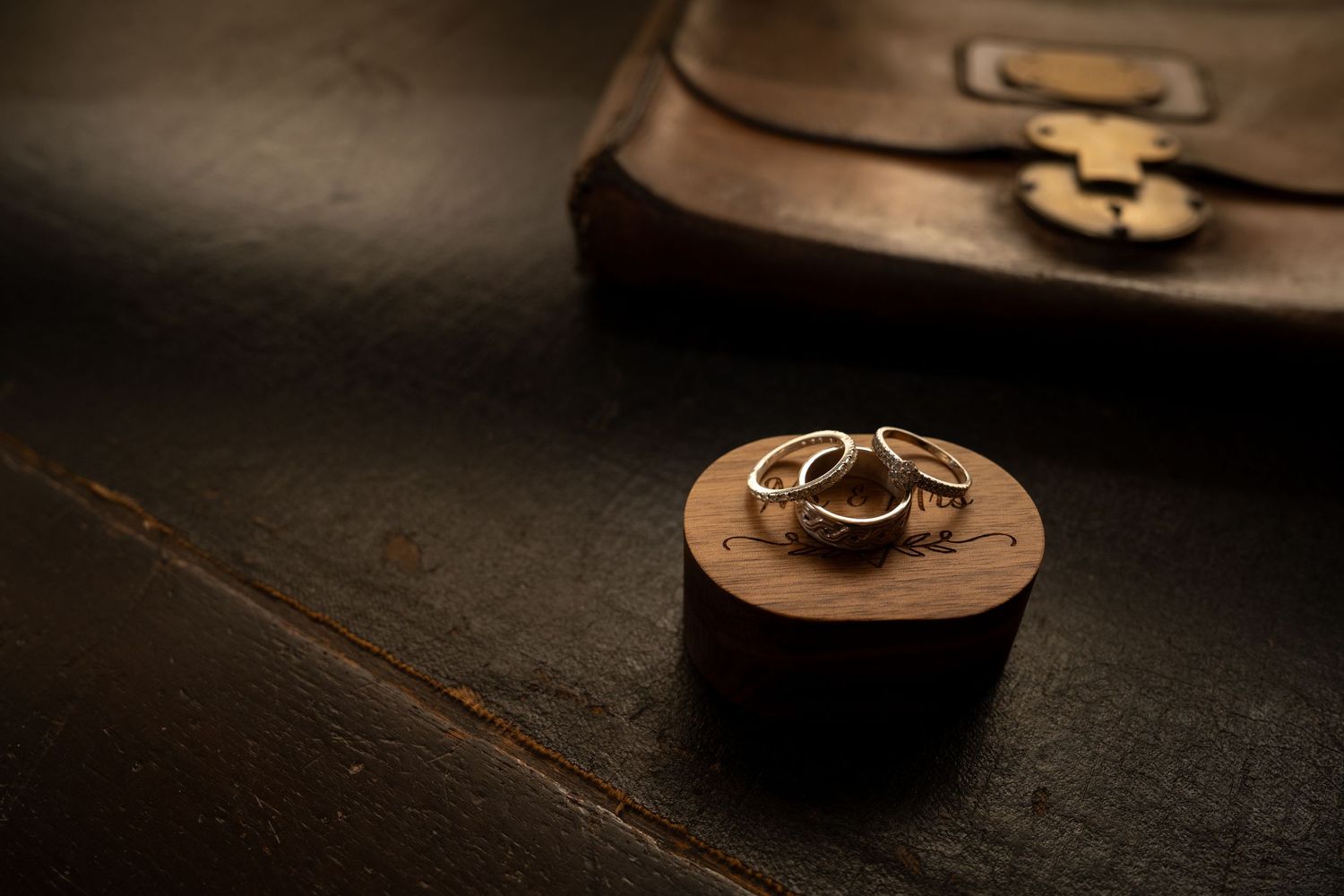 Two gold wedding rings rest on a round wooden holder in dramatic moody lighting with a leather bag in background.
