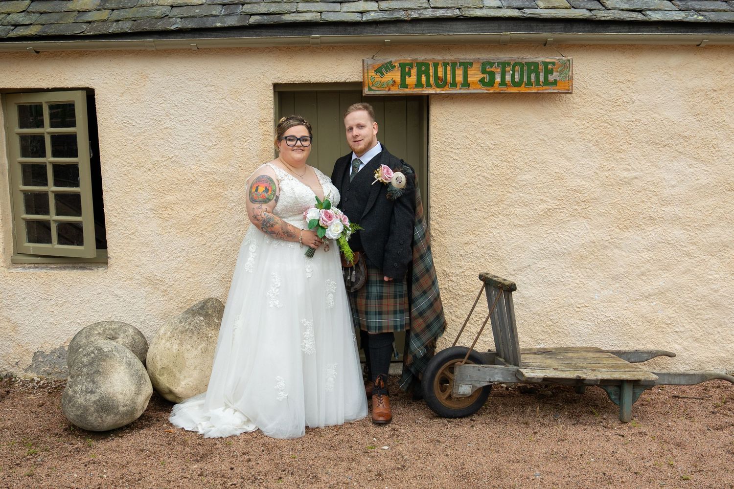 Rustic farmhouse wedding photoshoot with elegant white dress and traditional Scottish kilt against stone building wall.