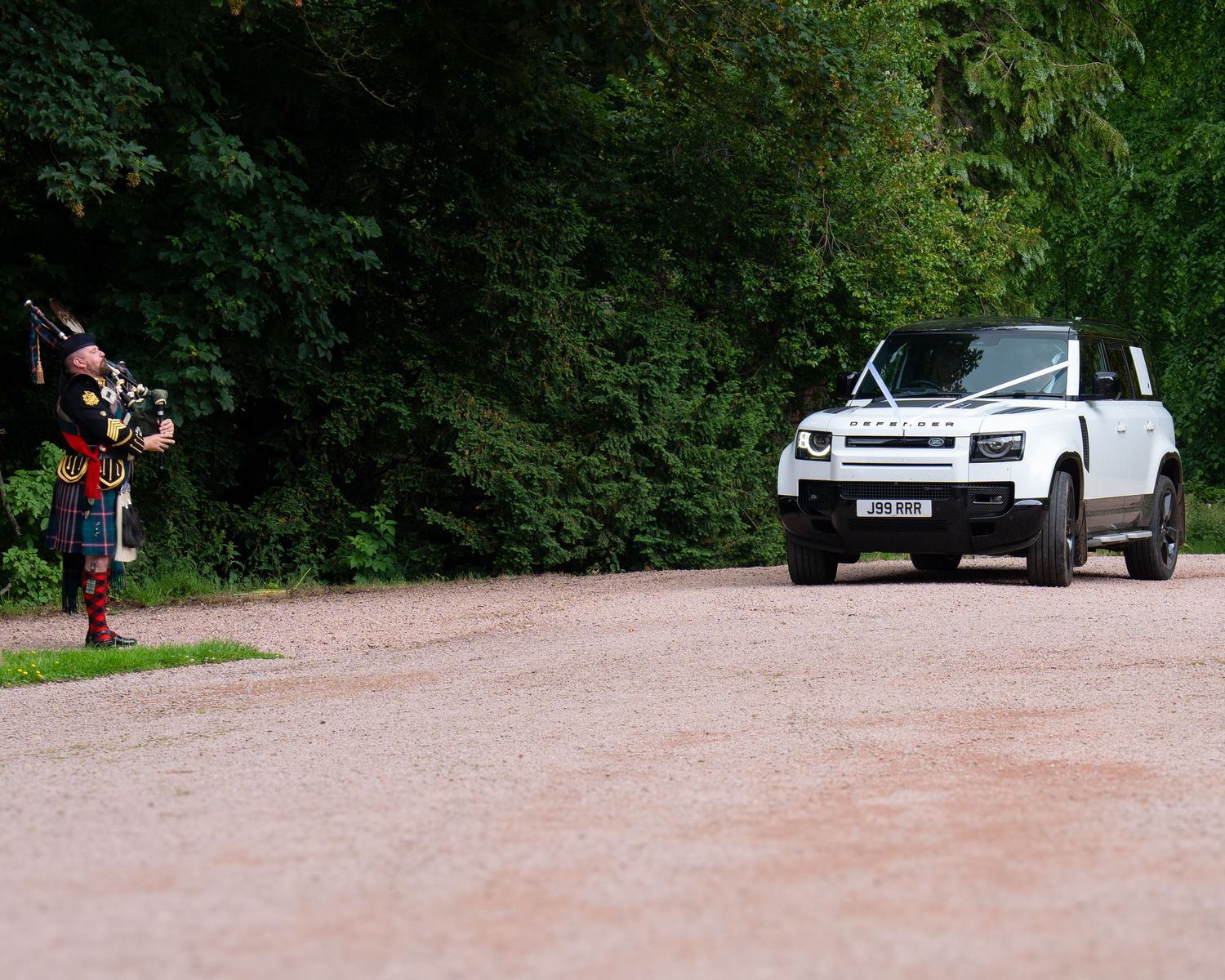 A white pickup truck on a dirt road while a bagpiper plays music in traditional Scottish attire.