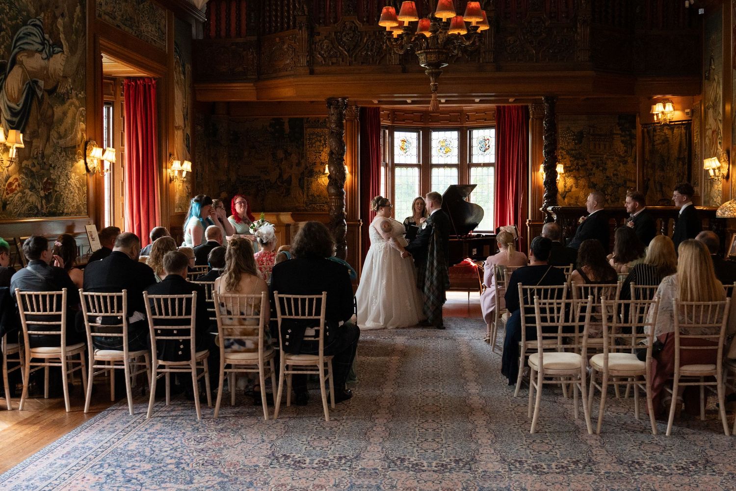 An elegant indoor wedding ceremony in a grand wood-paneled hall with chandeliers and white Chiavari chairs.