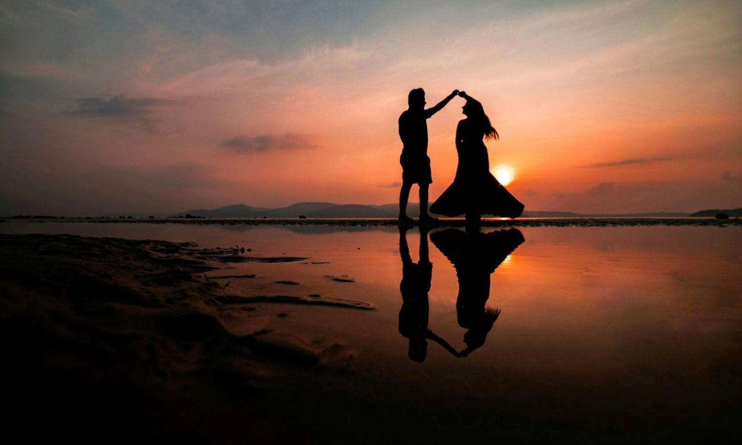 Silhouetted couple dancing together on a beach at sunset with dramatic reflections in the wet sand.