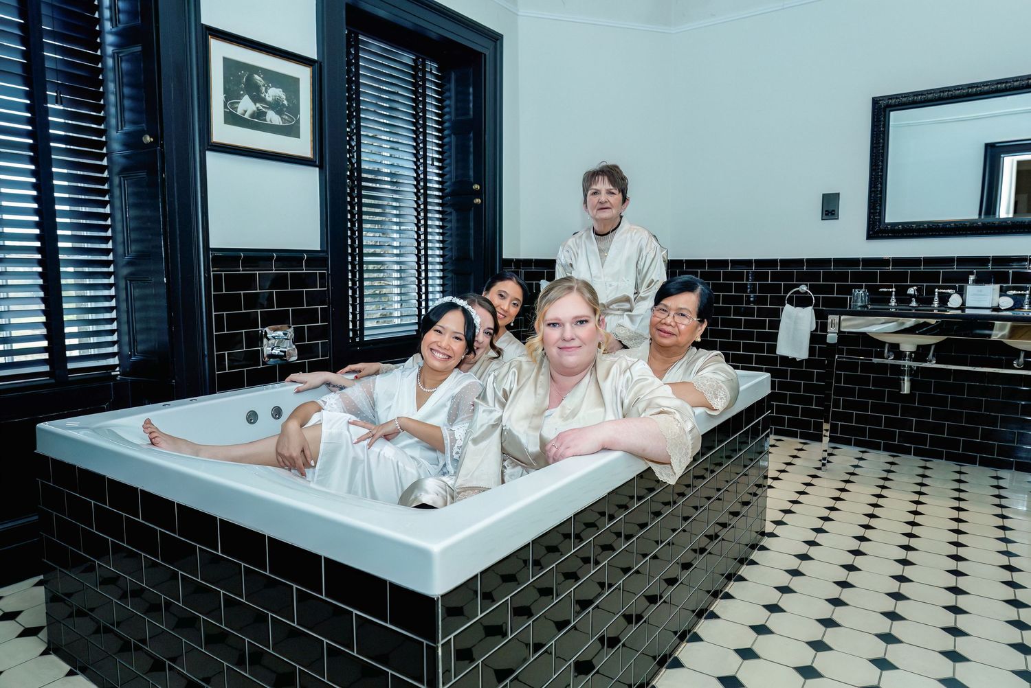 Relaxed newlyweds in white robes pose in vintage-style tiled bathroom with black and white checkerboard flooring.