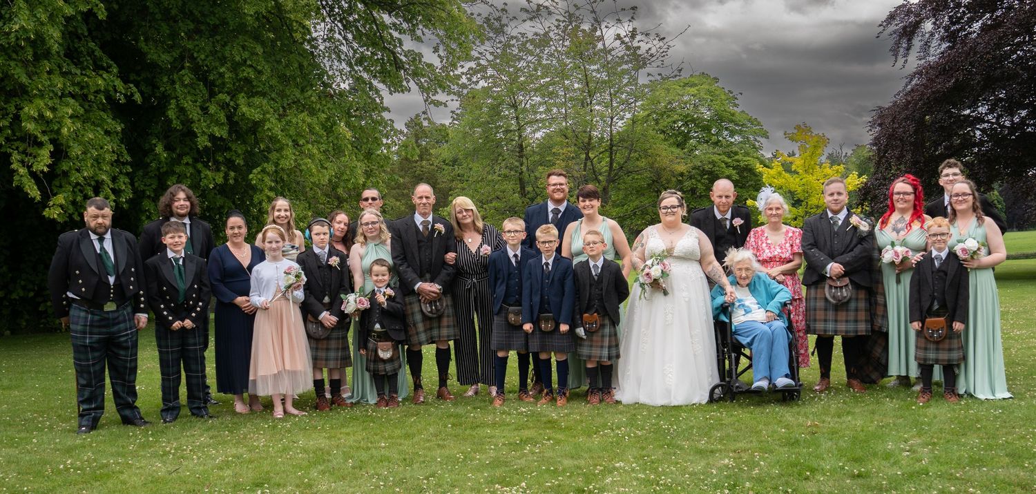 Wedding party members in traditional Scottish kilts and formal attire pose together on green lawn for group photo.