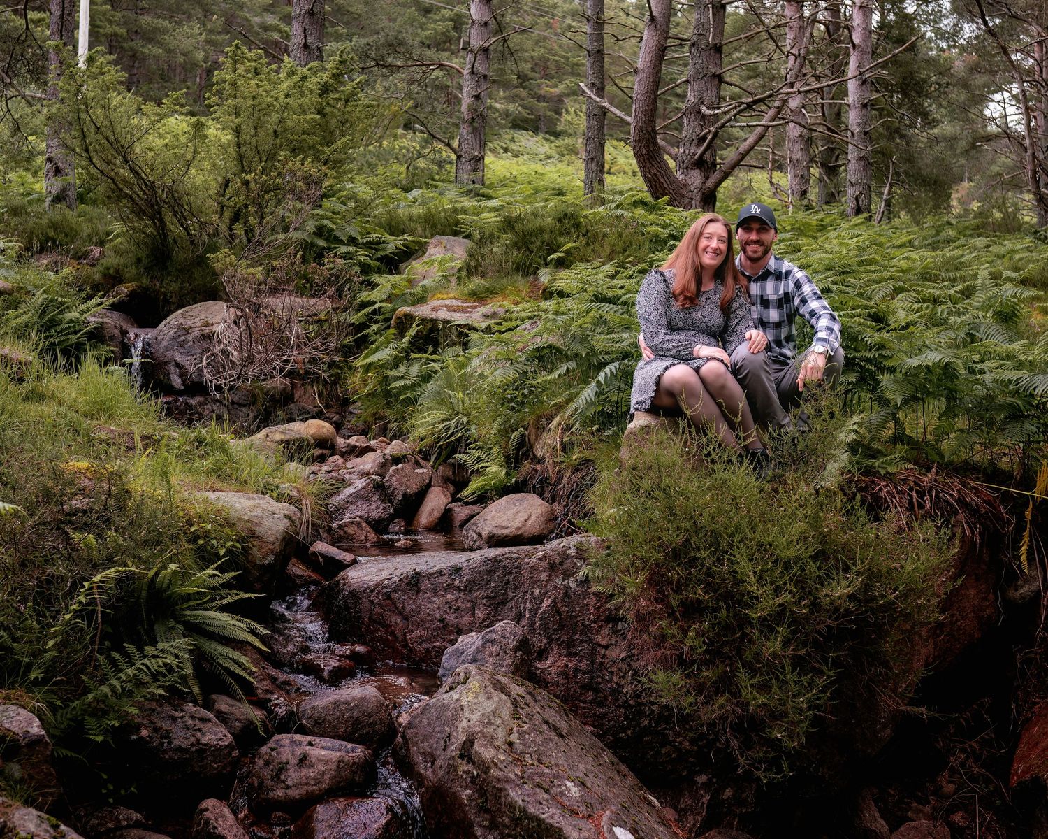 A couple sits together on mossy rocks beside a small forest stream surrounded by pine trees and ferns.
