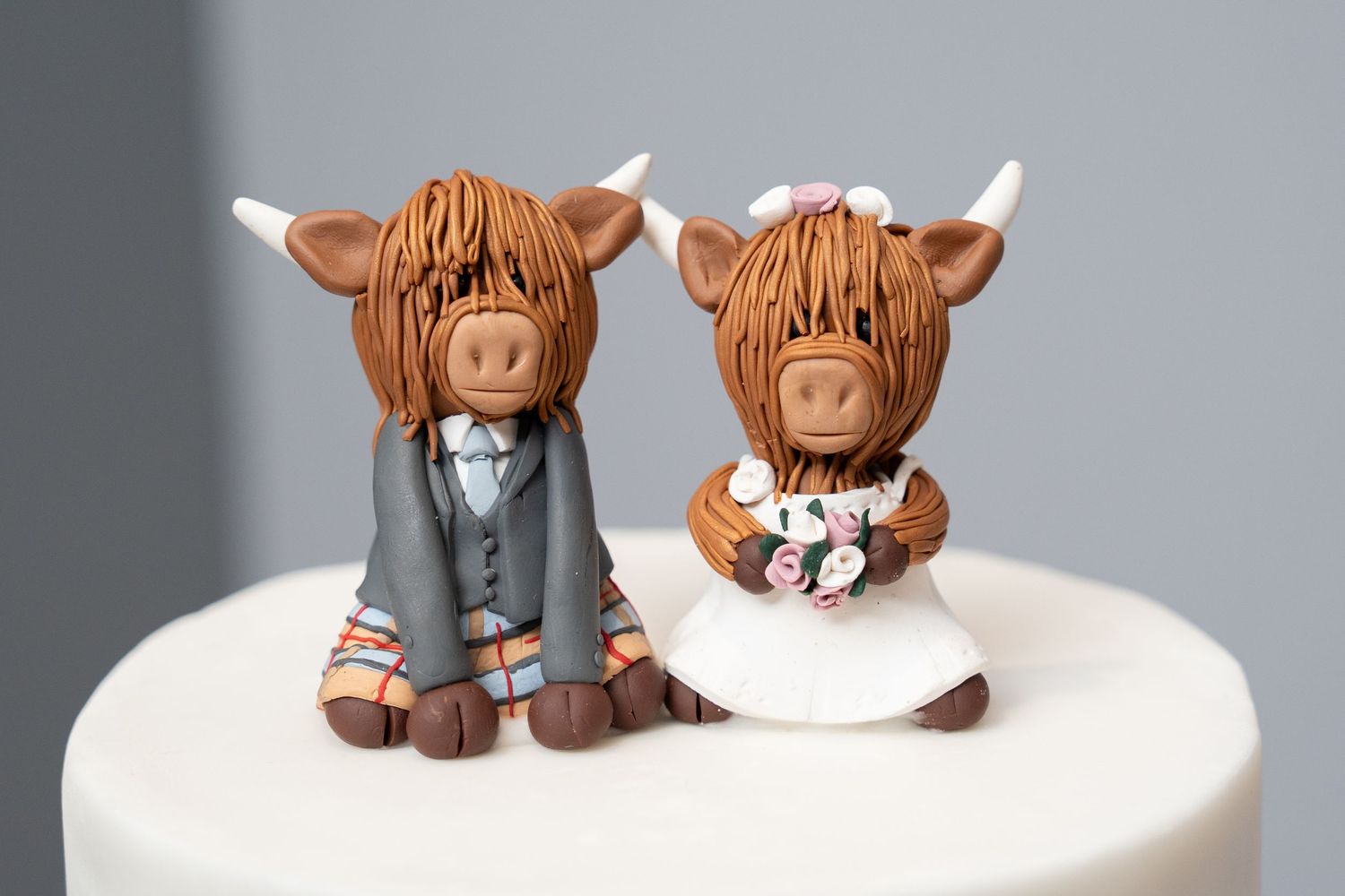 Adorable Highland cow bride and groom wedding cake toppers made from clay sit on a white cake.