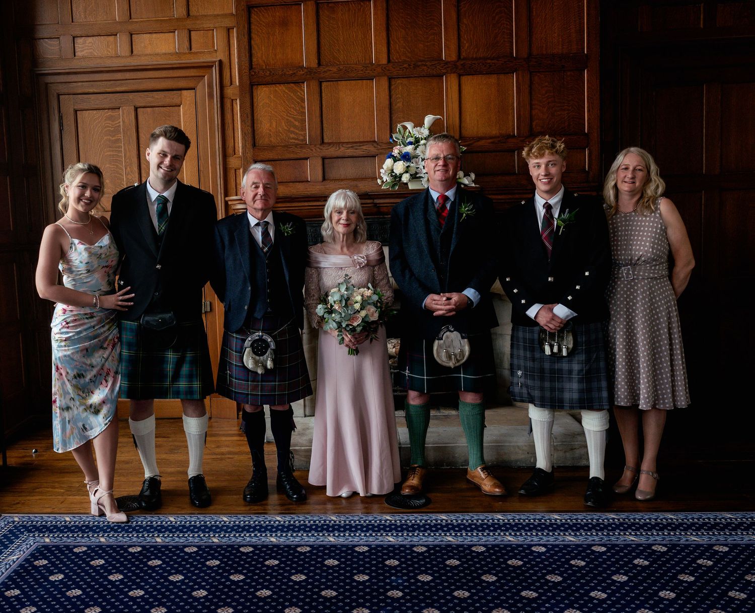 Wedding party group photo with men in kilts and formal attire standing together in a wood-paneled room.