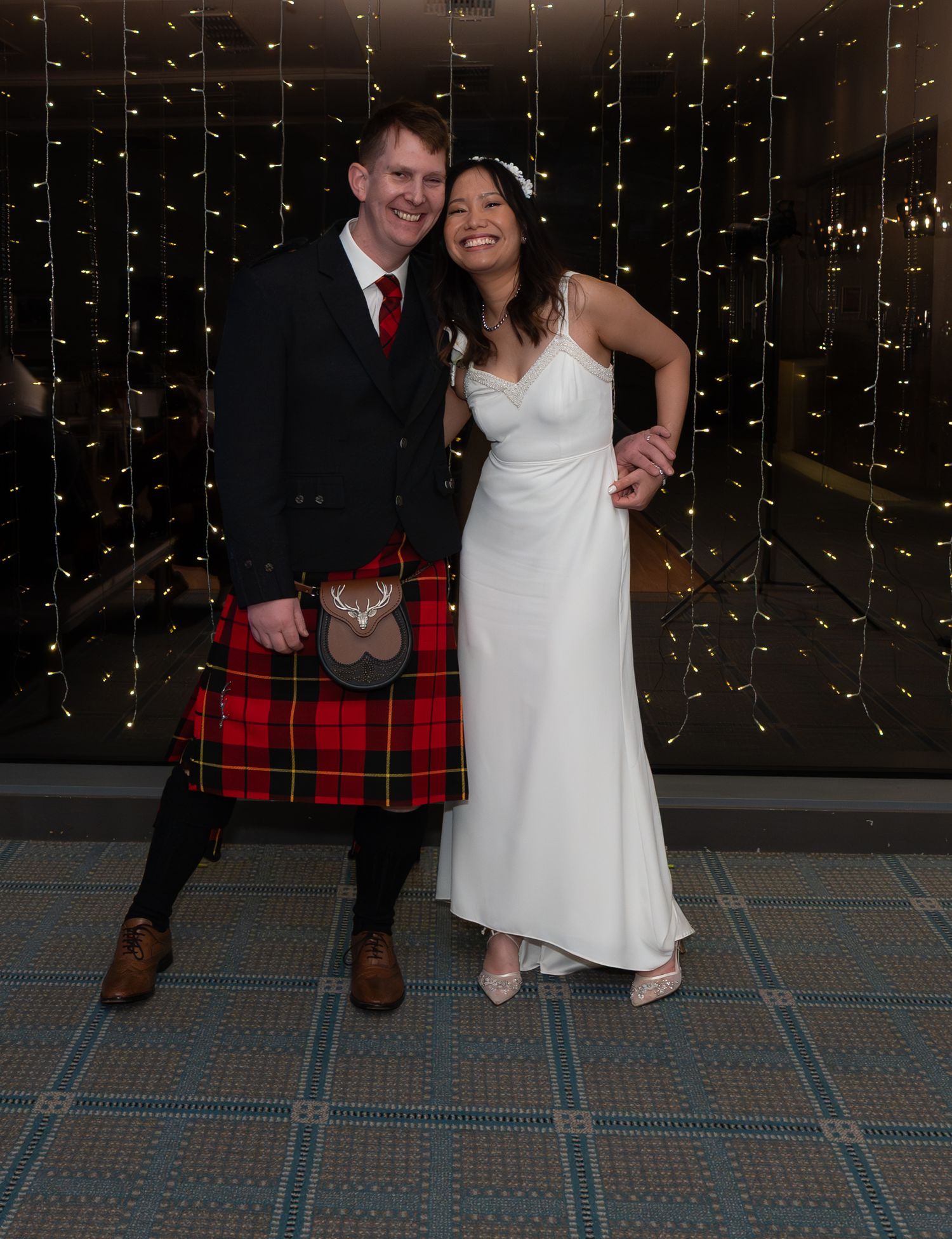 A couple poses at their wedding celebration, with the groom wearing a traditional red tartan kilt and the bride in a white gown.