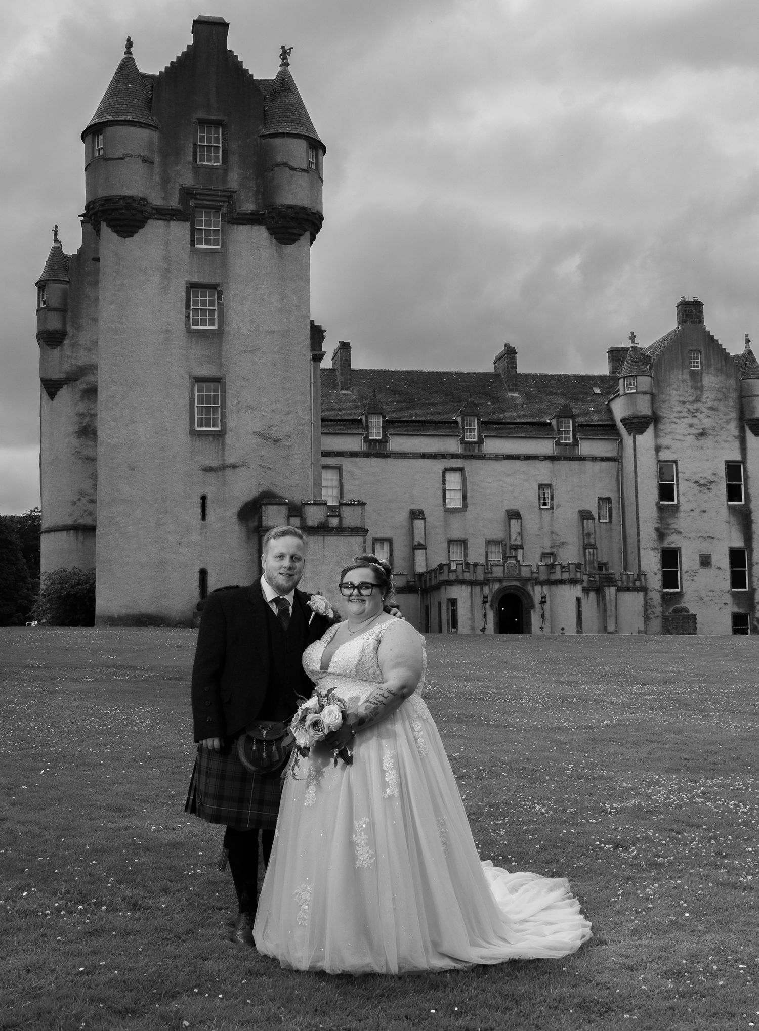 A wedding couple poses in front of a dramatic Scottish castle under moody gray skies.