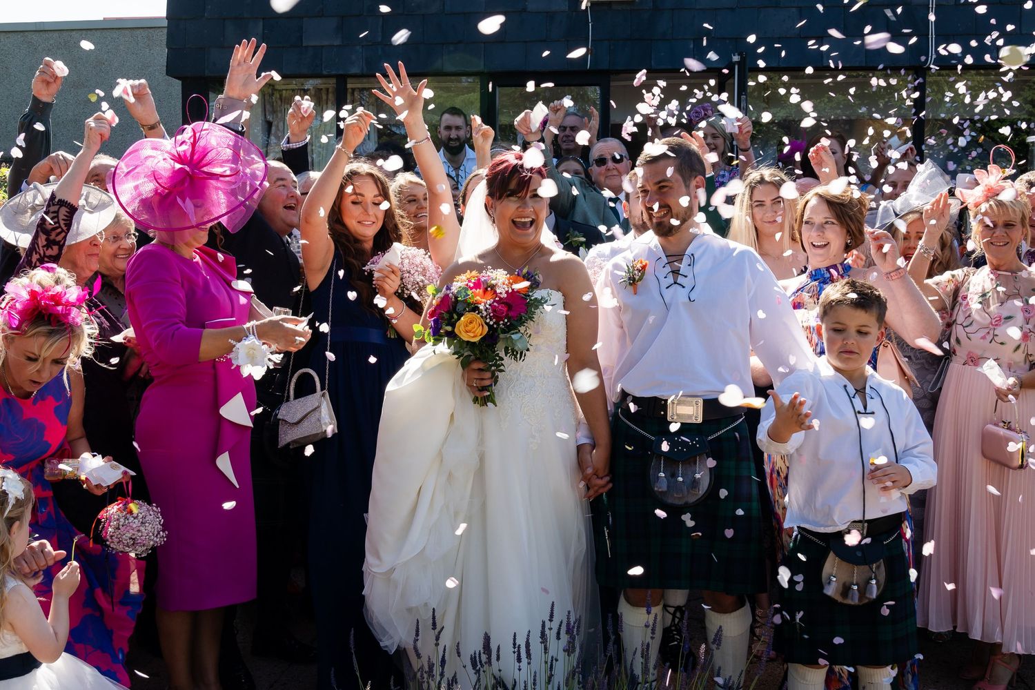 Large group of wedding guests tossing confetti in celebration outdoors.