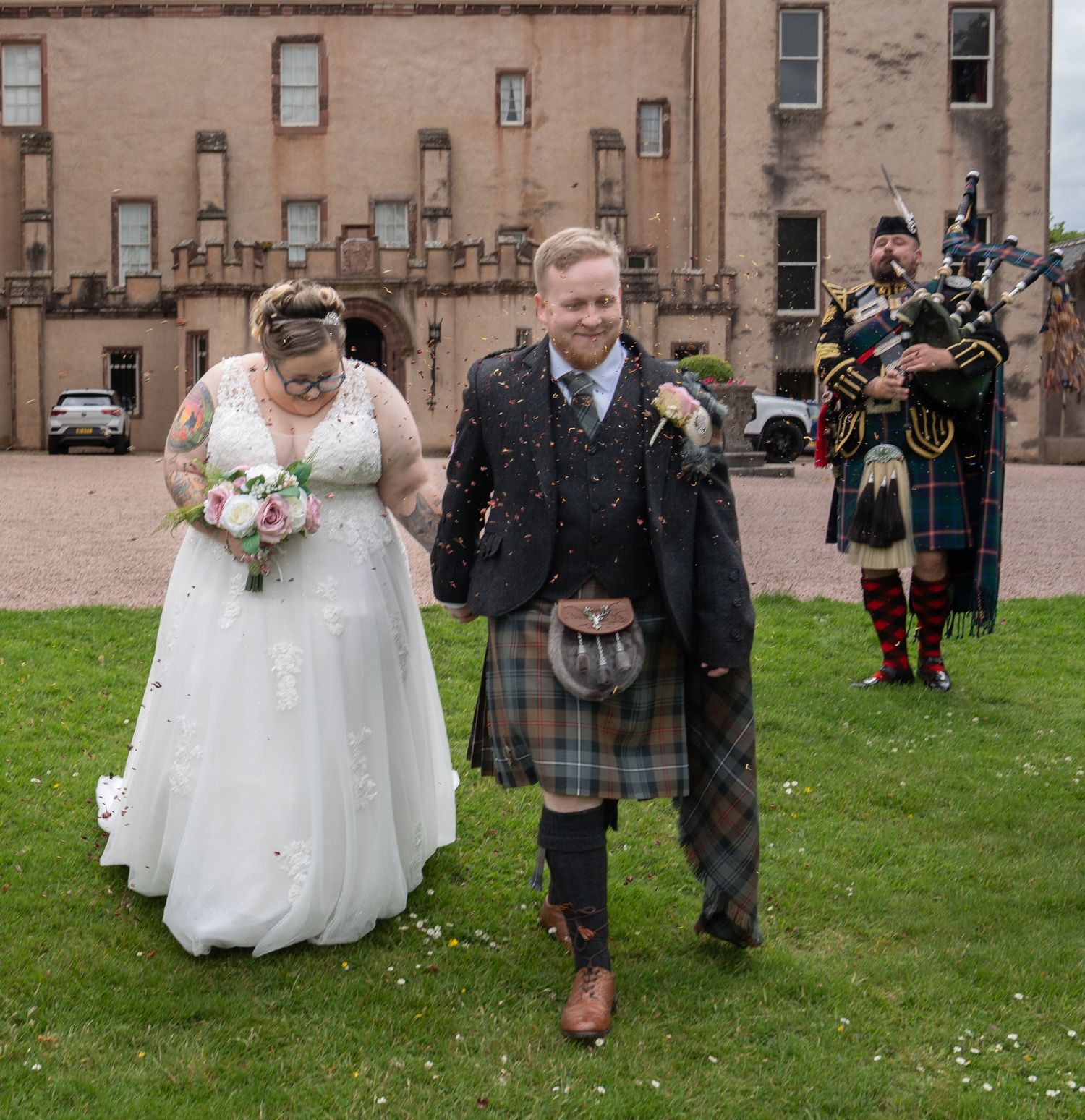 A traditional Scottish wedding ceremony with a bagpiper playing as couple walks on grass in front of stone building.