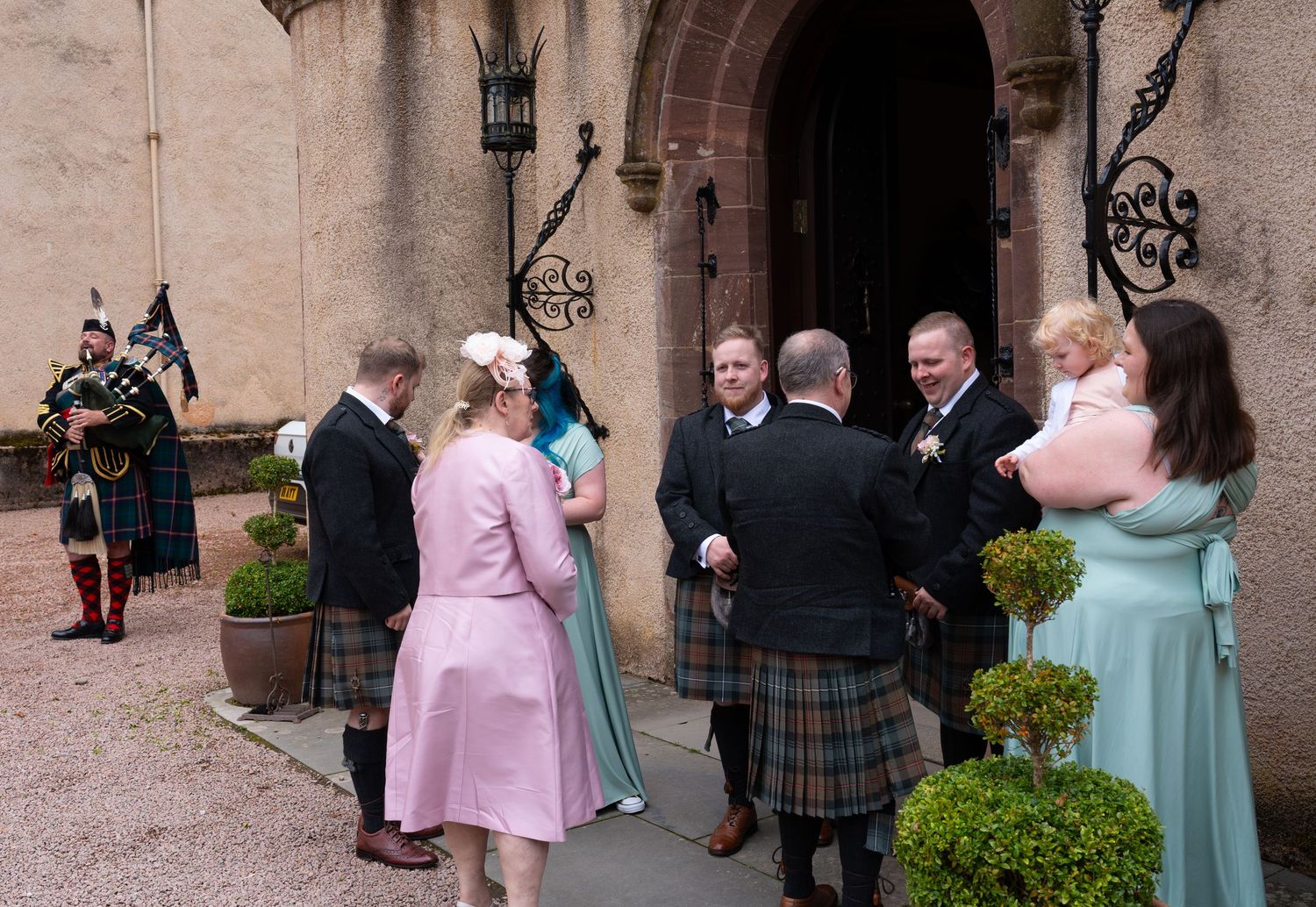 A group dressed in formal Scottish kilts and pastel dresses socializes outside a grand stone archway.