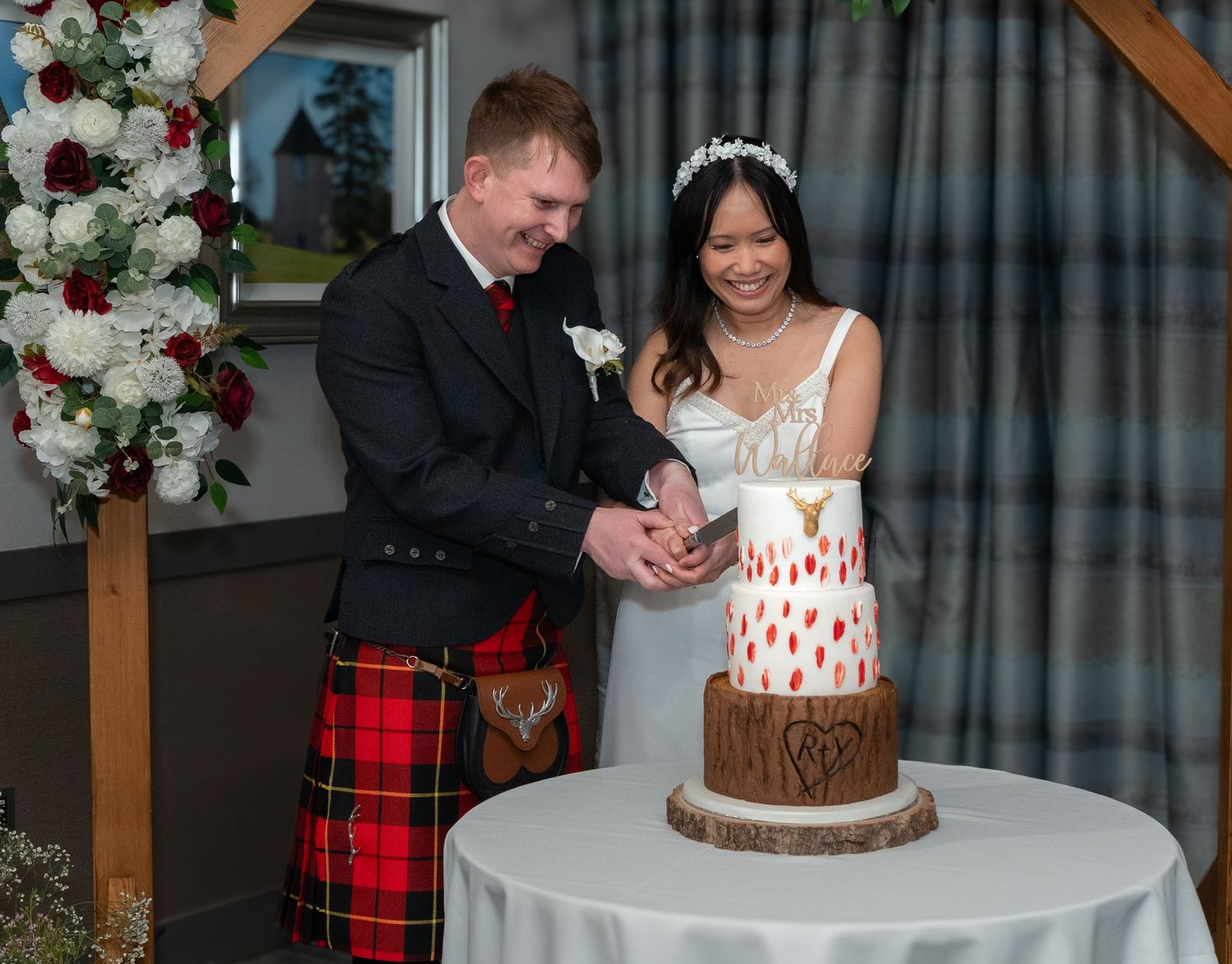 A newlywed couple cuts their rustic wooden wedding cake decorated with burgundy and white flowers beneath a wooden arbor.