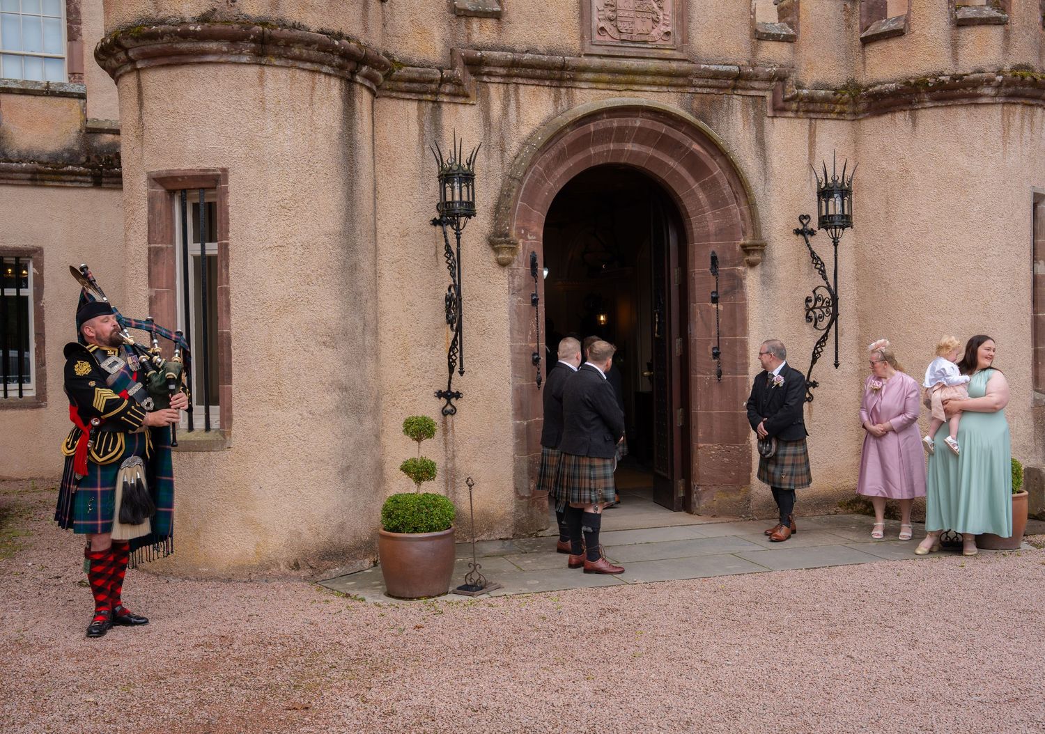 People in traditional Scottish attire and vintage dresses gather near an arched entrance of a historic stone building.