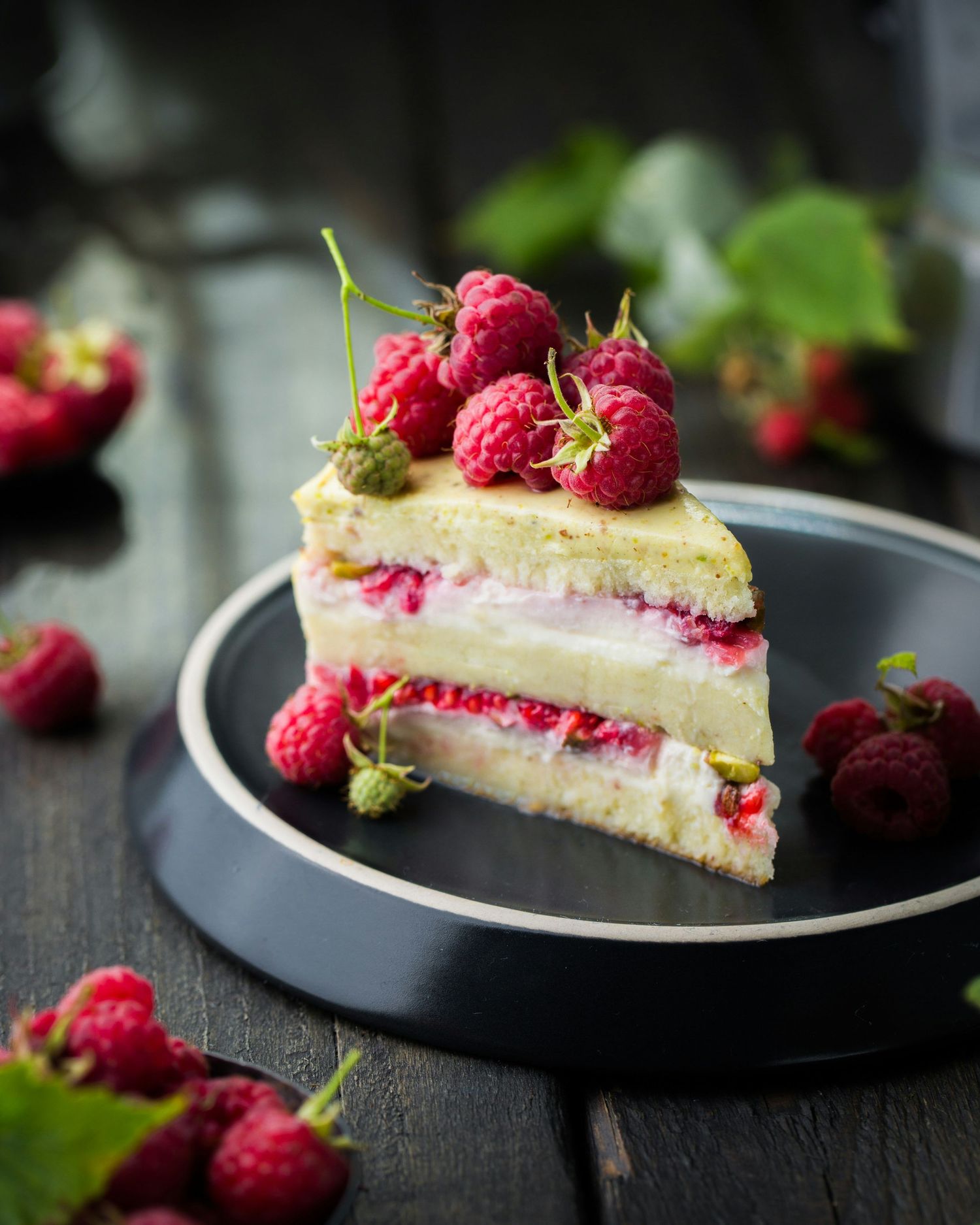 A slice of layered vanilla cake with fresh raspberries and cream filling on a gray plate against a dark wood background.