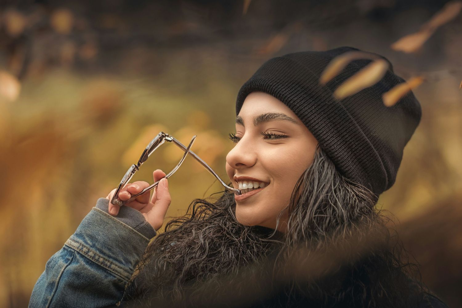 A person wearing a black beanie and denim jacket smiles while holding glasses on an autumn day.
