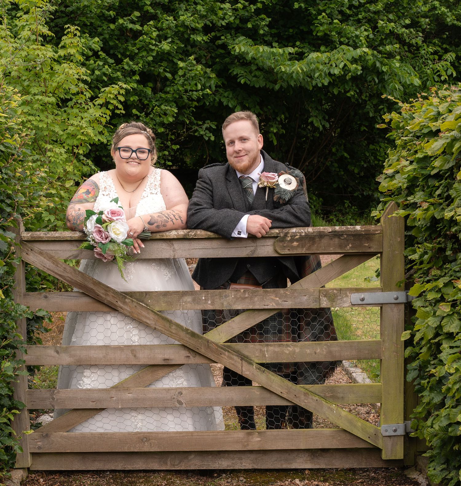 A couple leans on a rustic wooden gate surrounded by lush greenery.
