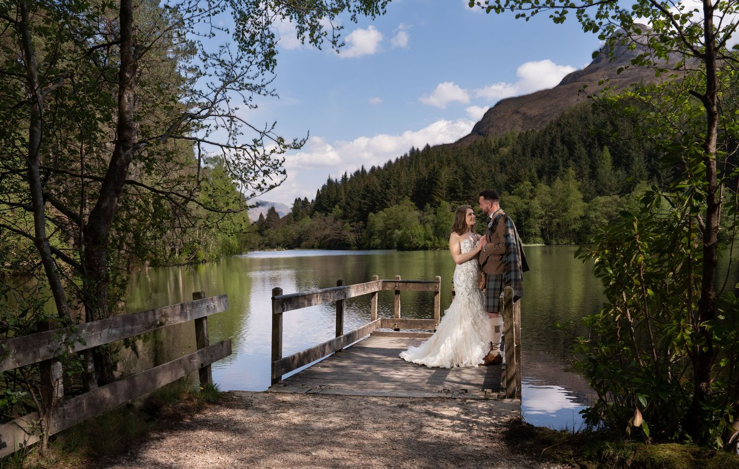 A romantic wedding moment captured on a wooden bridge over a tranquil lake surrounded by mountains.