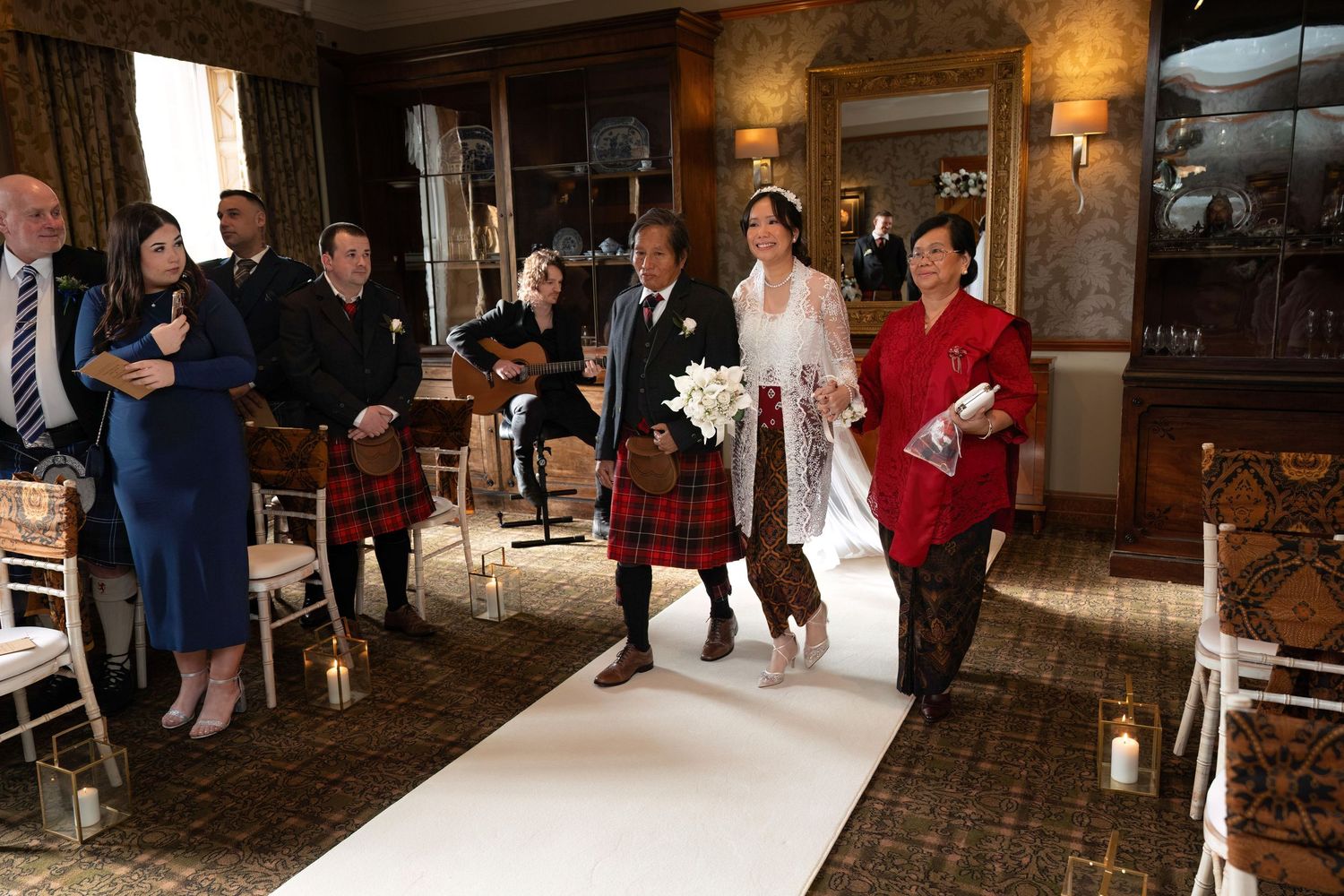 Wedding ceremony in a rustic venue with traditional Scottish kilts and formal attire as guests gather along the aisle.