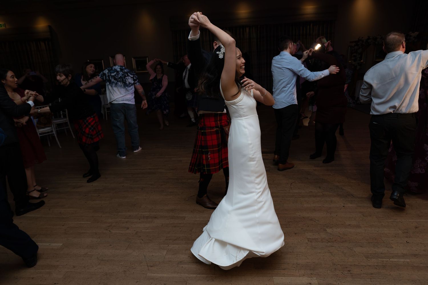 A person in a white wedding dress twirls and dances on a wooden dance floor at a celebration with others in the background.