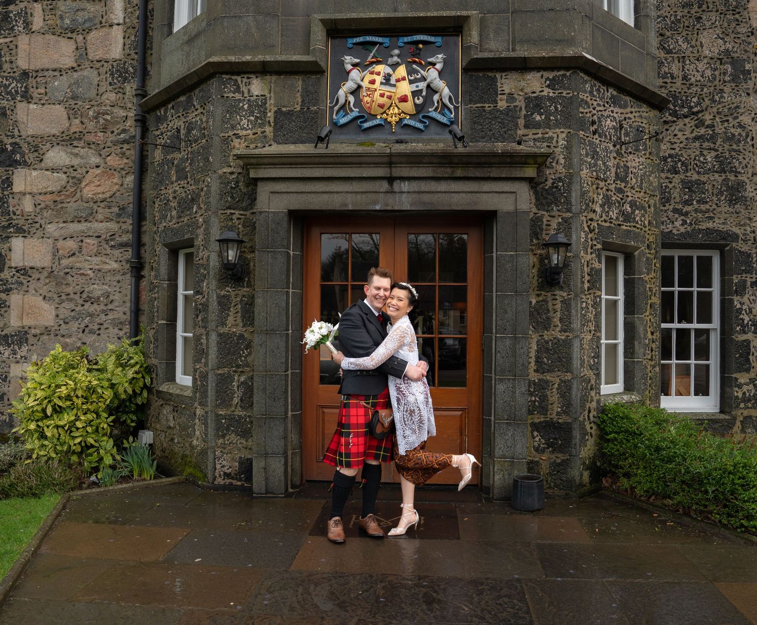 A couple embraces in front of a grand stone entrance with a heraldic crest above wooden doors at a Scottish estate.
