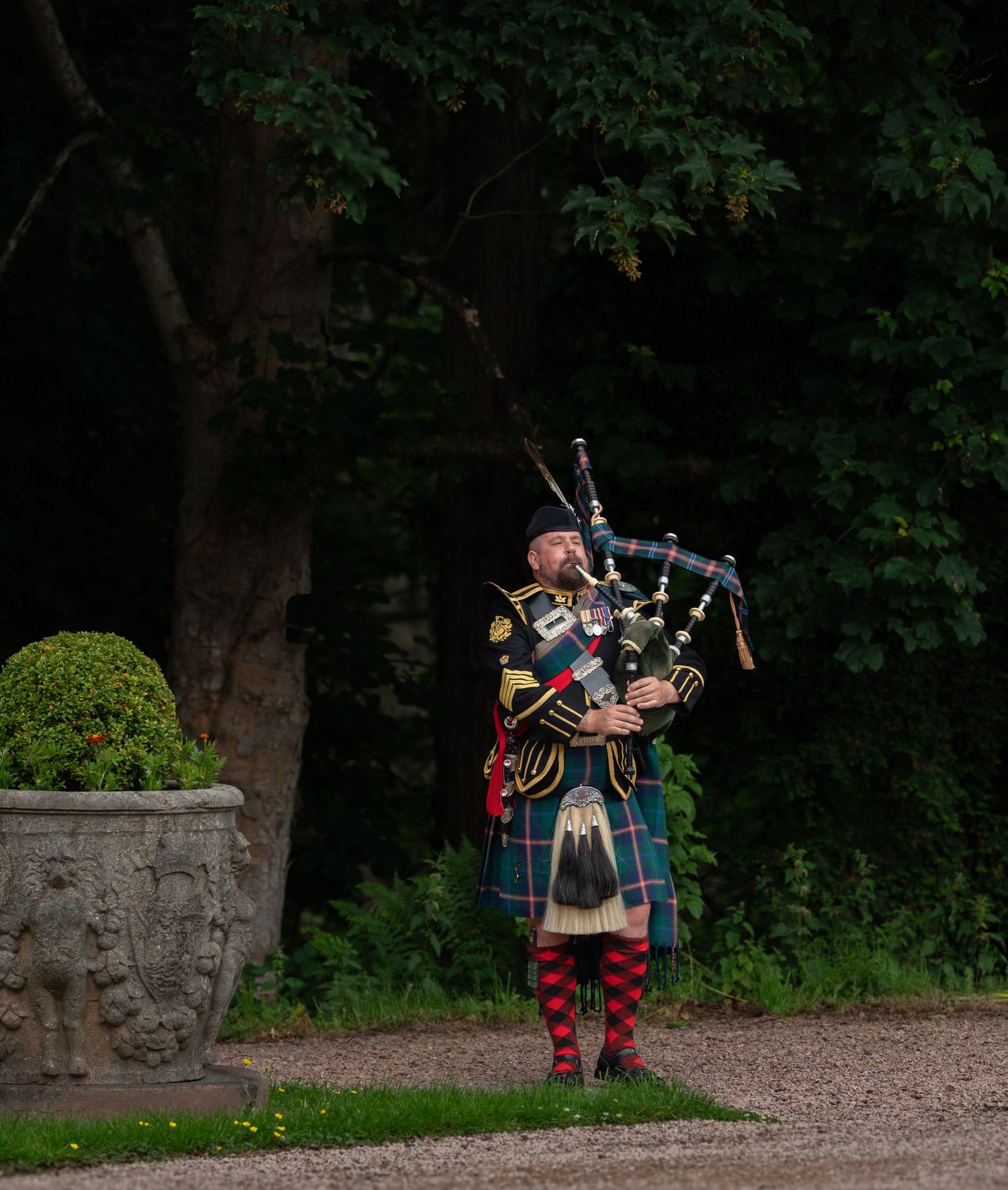 A sequence of bagpipe players performing in traditional Scottish attire on a gravel path near a stone wall and trees.