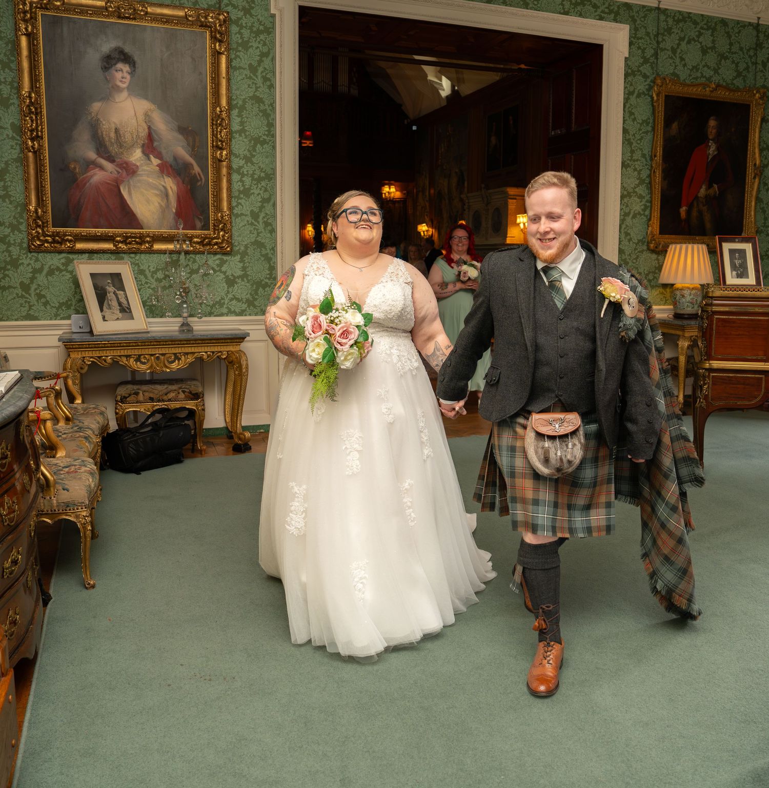 A wedding couple stands in an elegant room with green wallpaper and ornate gold-framed artwork, the groom wearing a traditional kilt.