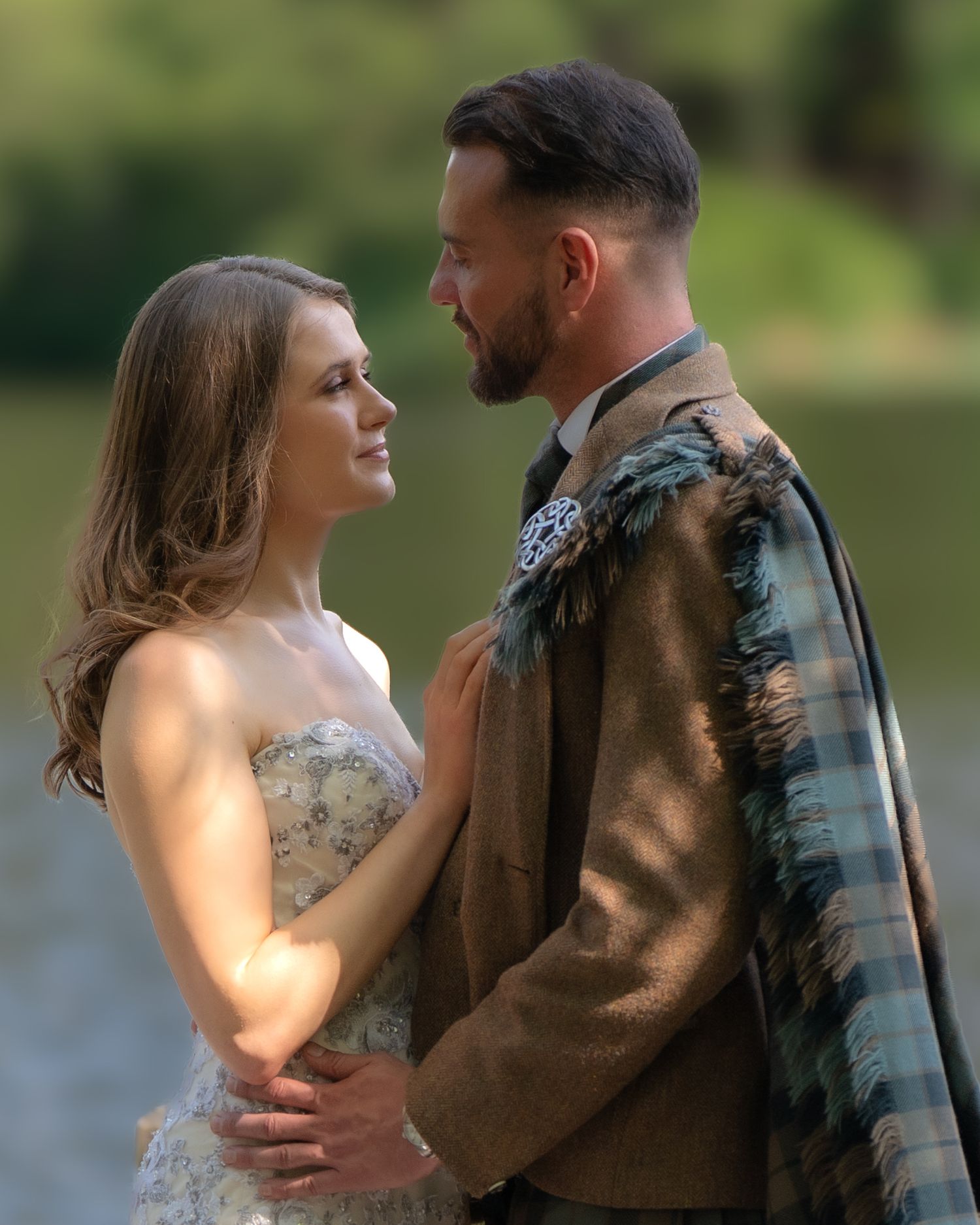 A couple in wedding attire embraces outdoors, with the bride in a lace dress and groom in a casual brown jacket.