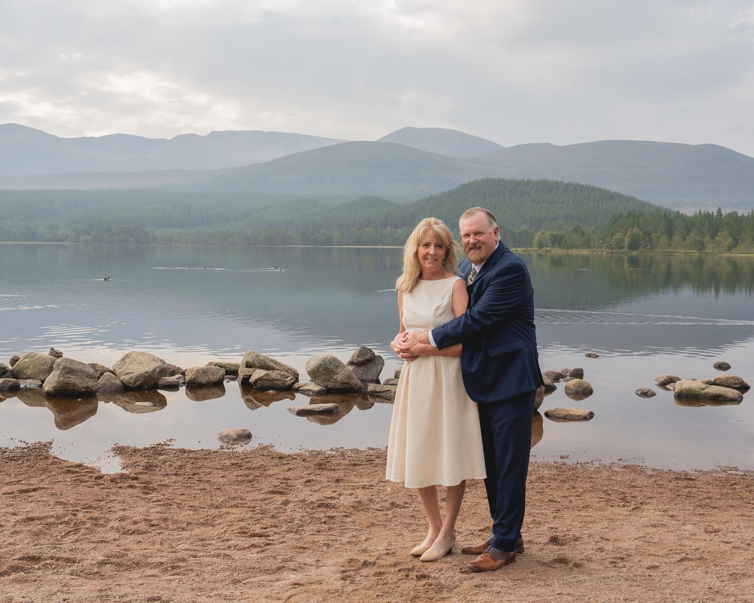 A couple stands together on a beach with mountains and a serene lake in the background.