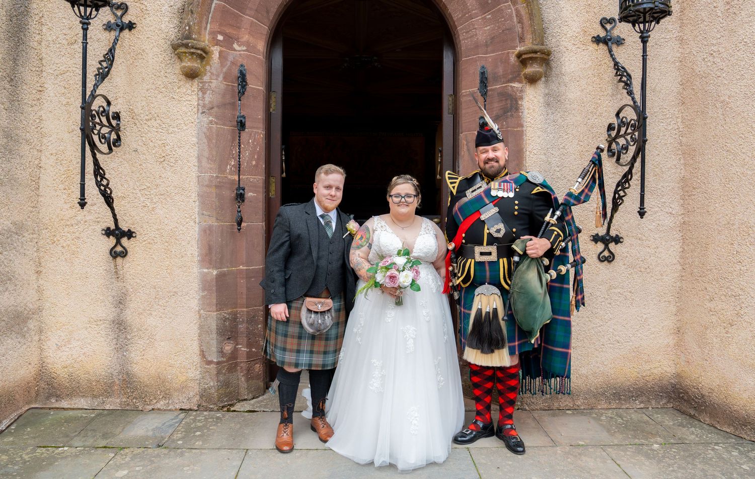 A wedding portrait at a stone church entrance with traditional Scottish attire and kilts.