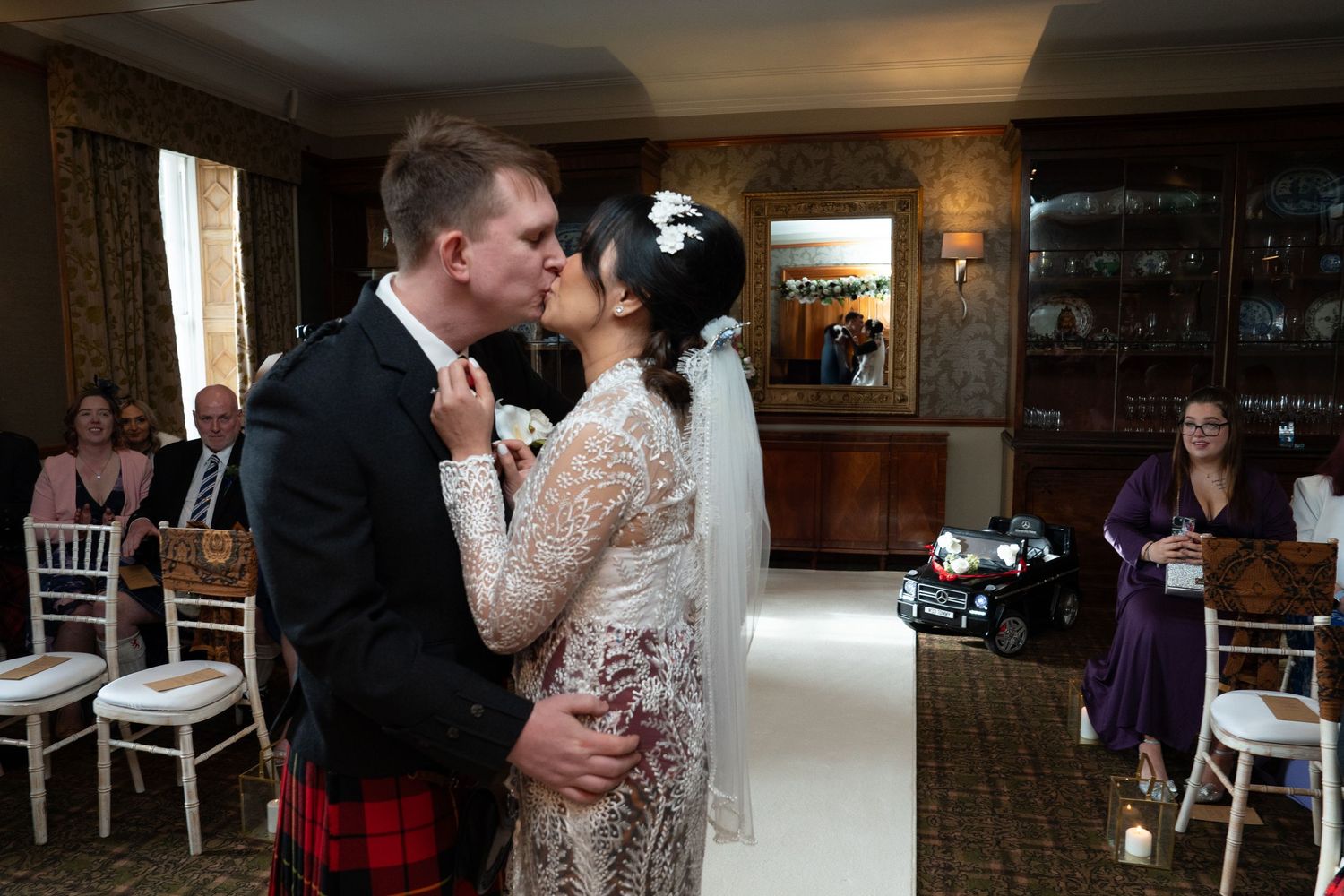 A couple shares their first dance at a wedding reception in a rustic room with wood paneling and white chairs.