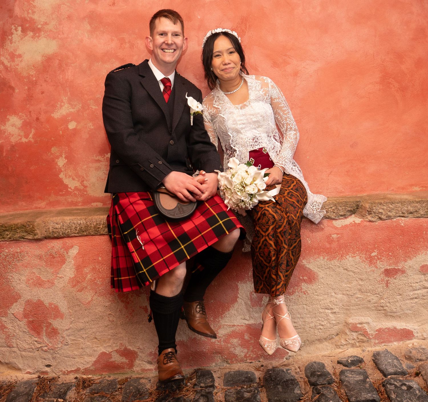 Wedding couple poses against rustic coral-colored wall in traditional Scottish and Indonesian wedding attire.