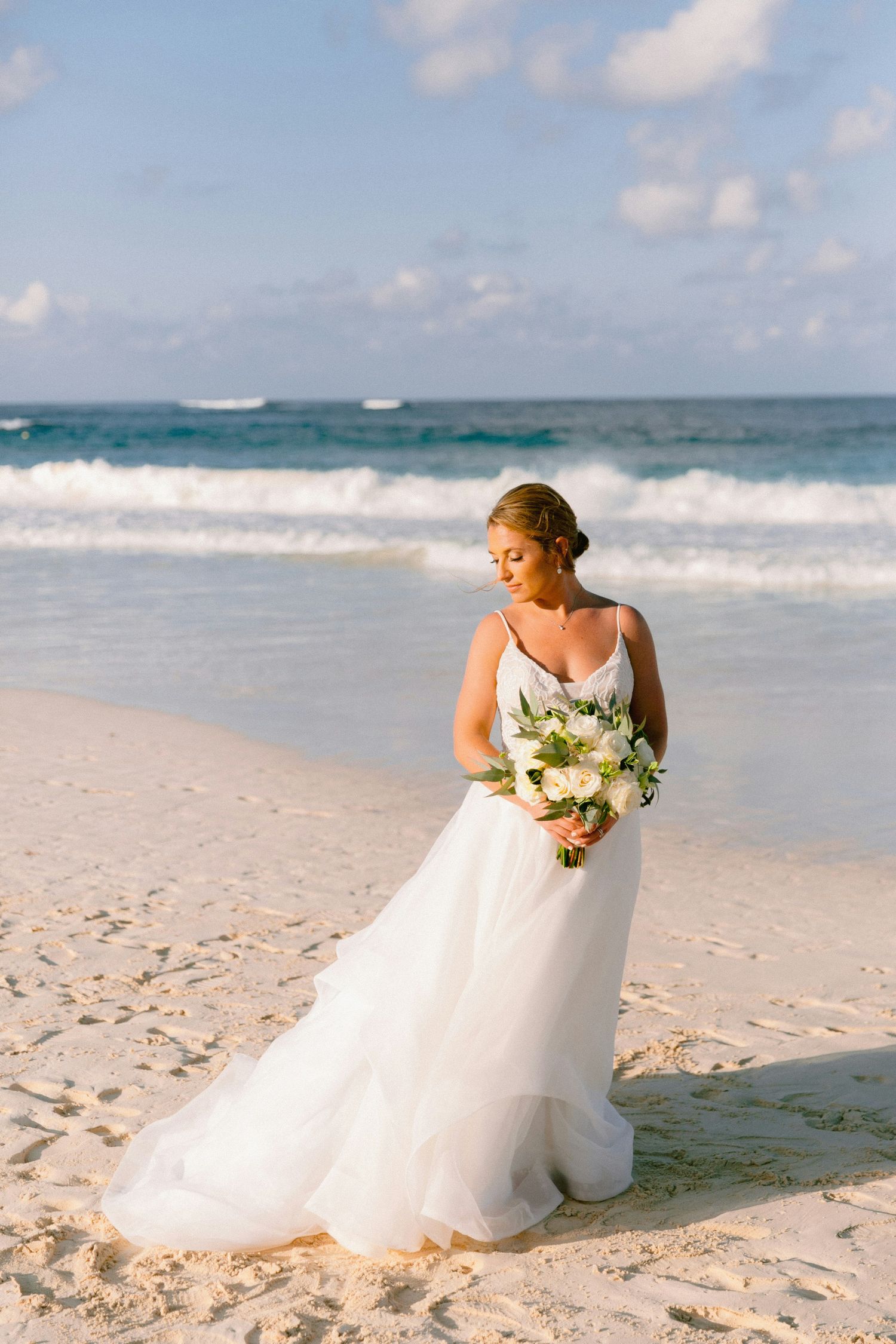 Bride in flowing white wedding gown poses gracefully along pristine beach with ocean waves and blue sky in background.
