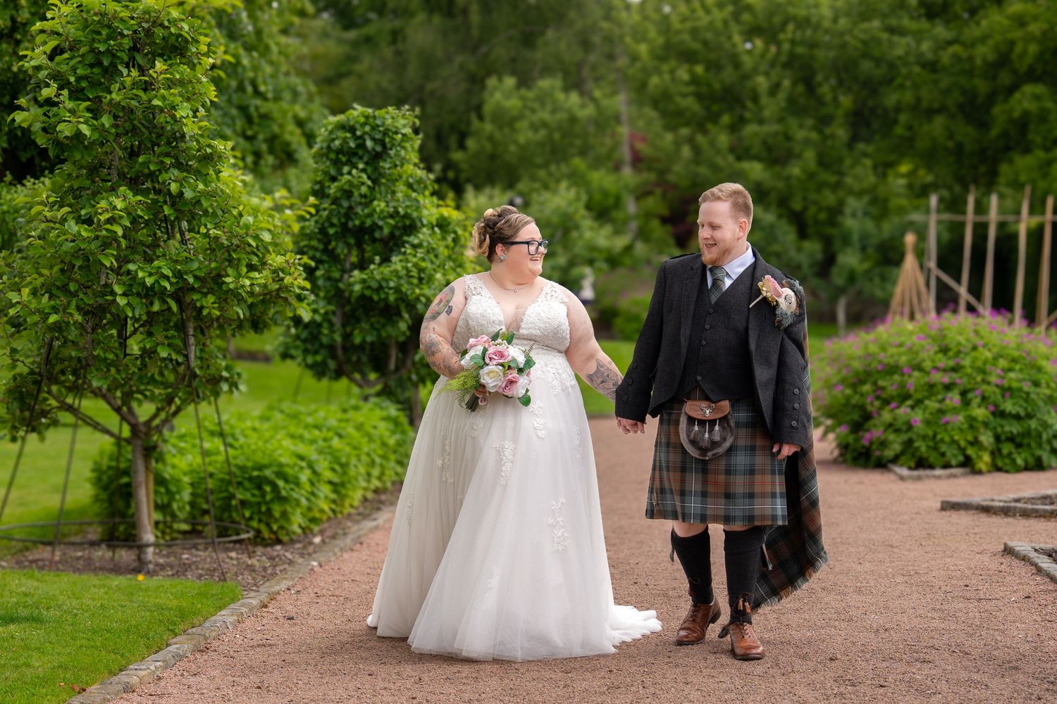 A couple stands in a scenic garden path during their wedding celebration, surrounded by lush greenery and trees.