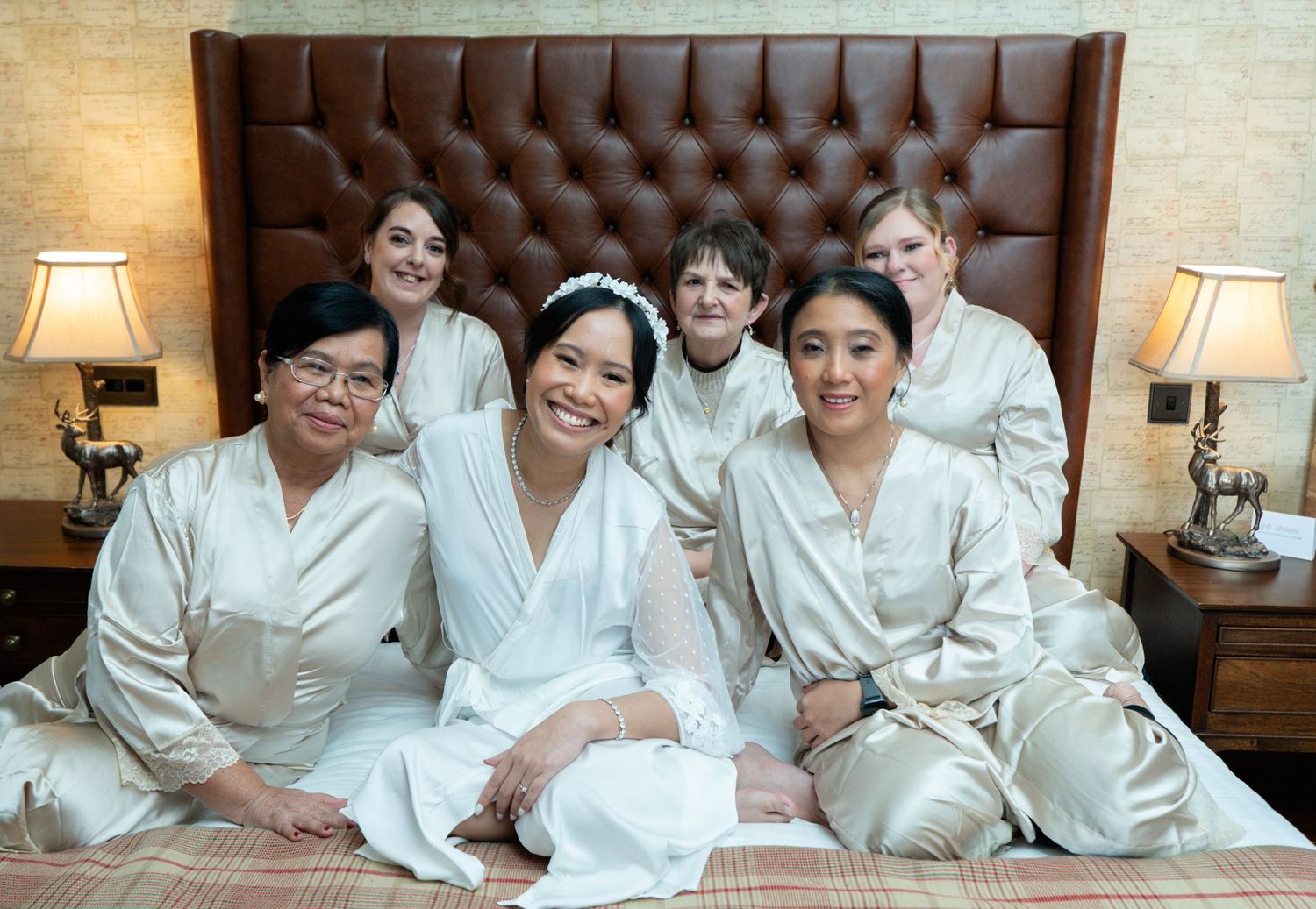 A group of seven people wearing matching white robes sit together on a bed with a brown tufted headboard and matching lamps.