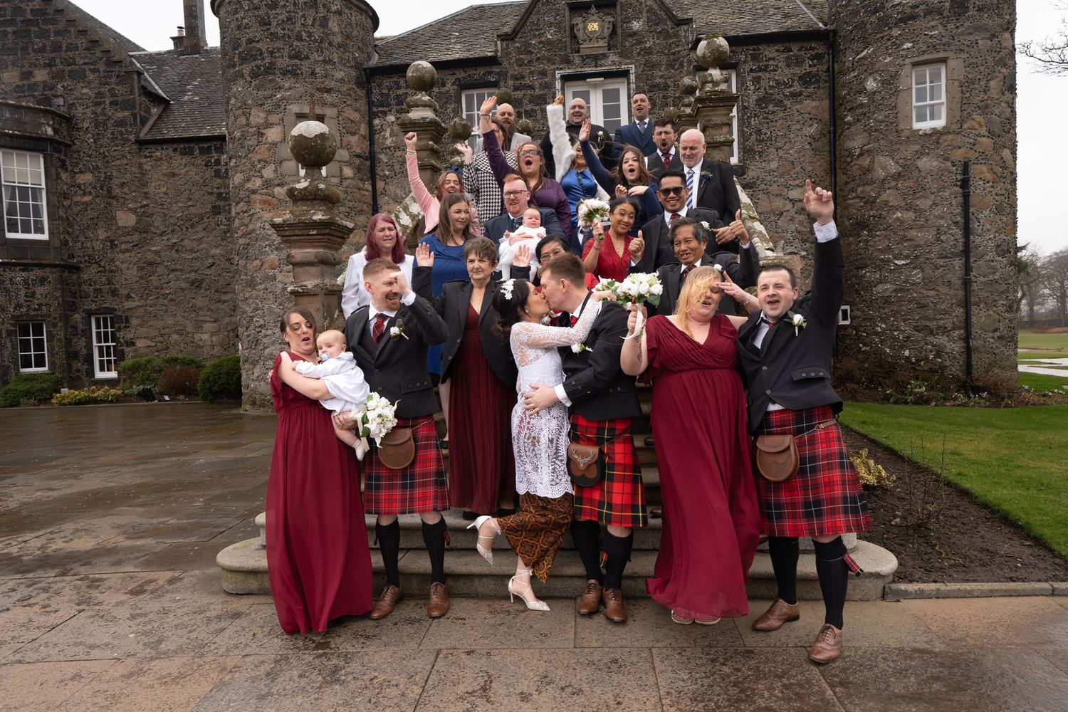 Wedding party celebrating outside a historic stone castle in Scotland with traditional kilts and burgundy dresses.