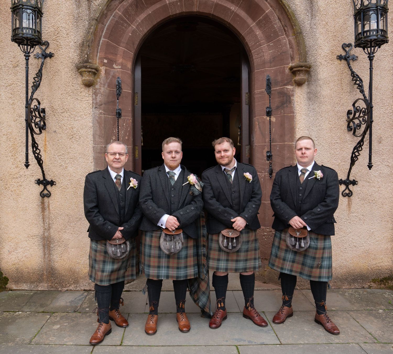 Four groomsmen wearing traditional Scottish kilts and formal jackets pose in front of an arched stone entrance.
