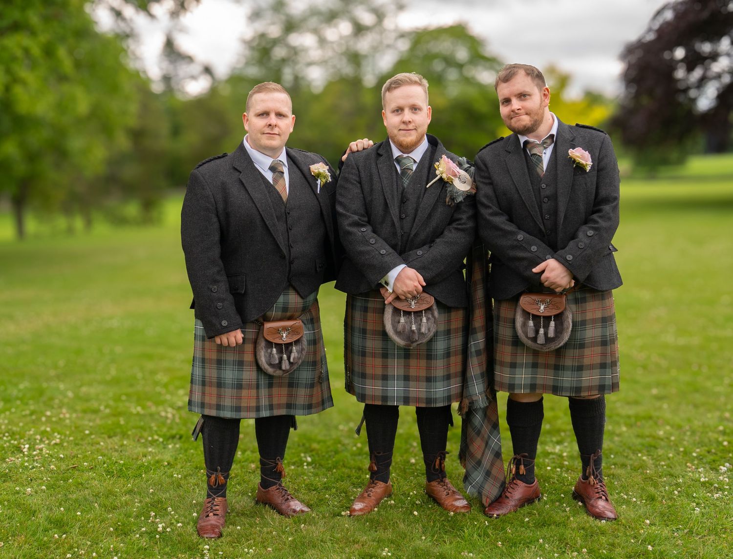 Three groomsmen wearing traditional Scottish kilts and black jackets pose together on green grass at a wedding.