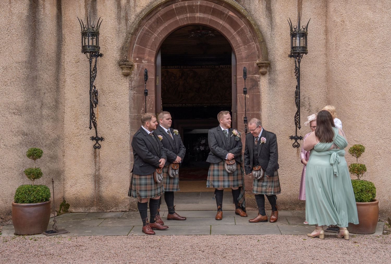 People in traditional Scottish kilts gather outside a stone church entrance with decorative lanterns and potted plants.