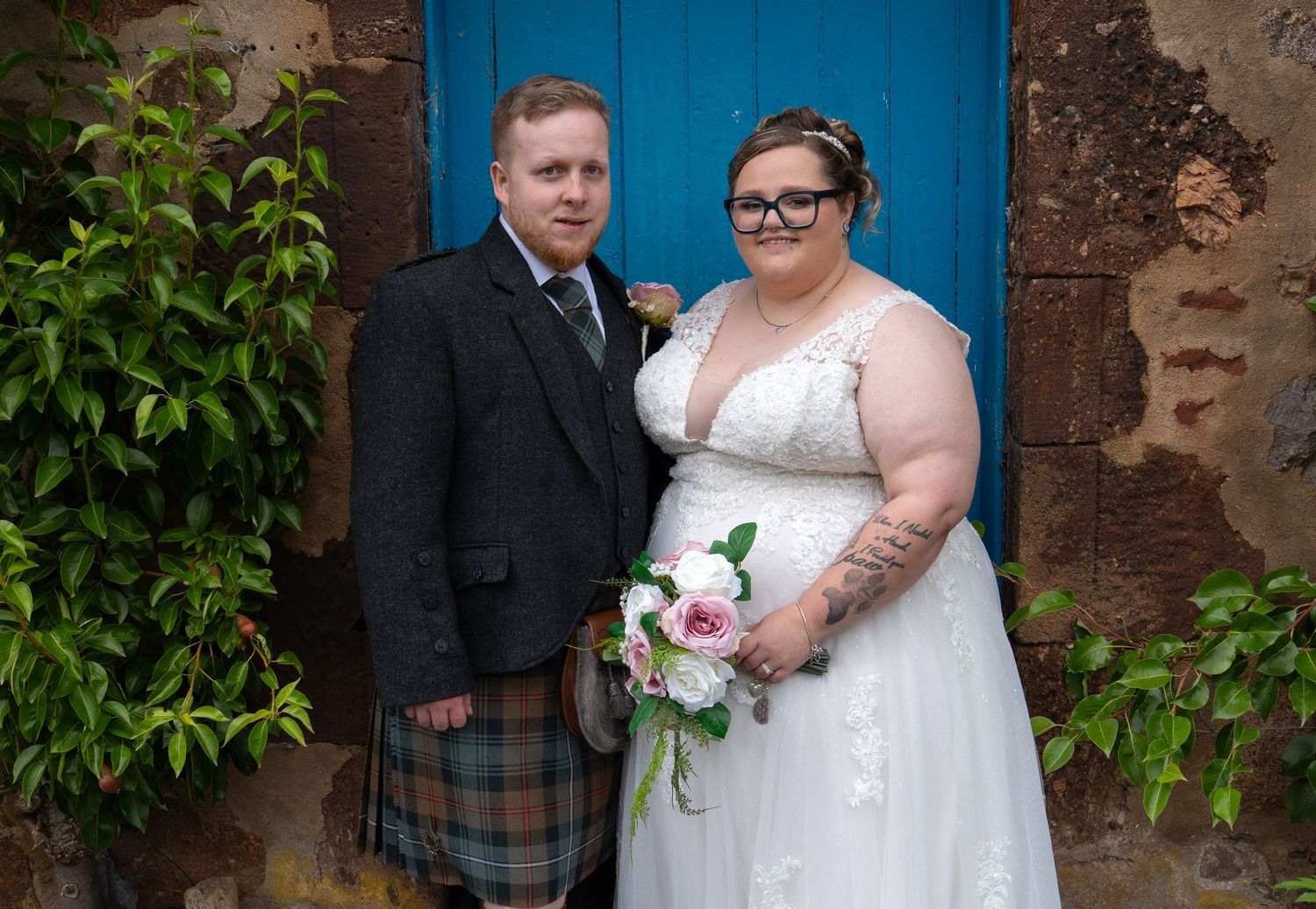 A couple stands together at their Scottish wedding, with the groom in a kilt and bride in white holding a bouquet by a blue door.