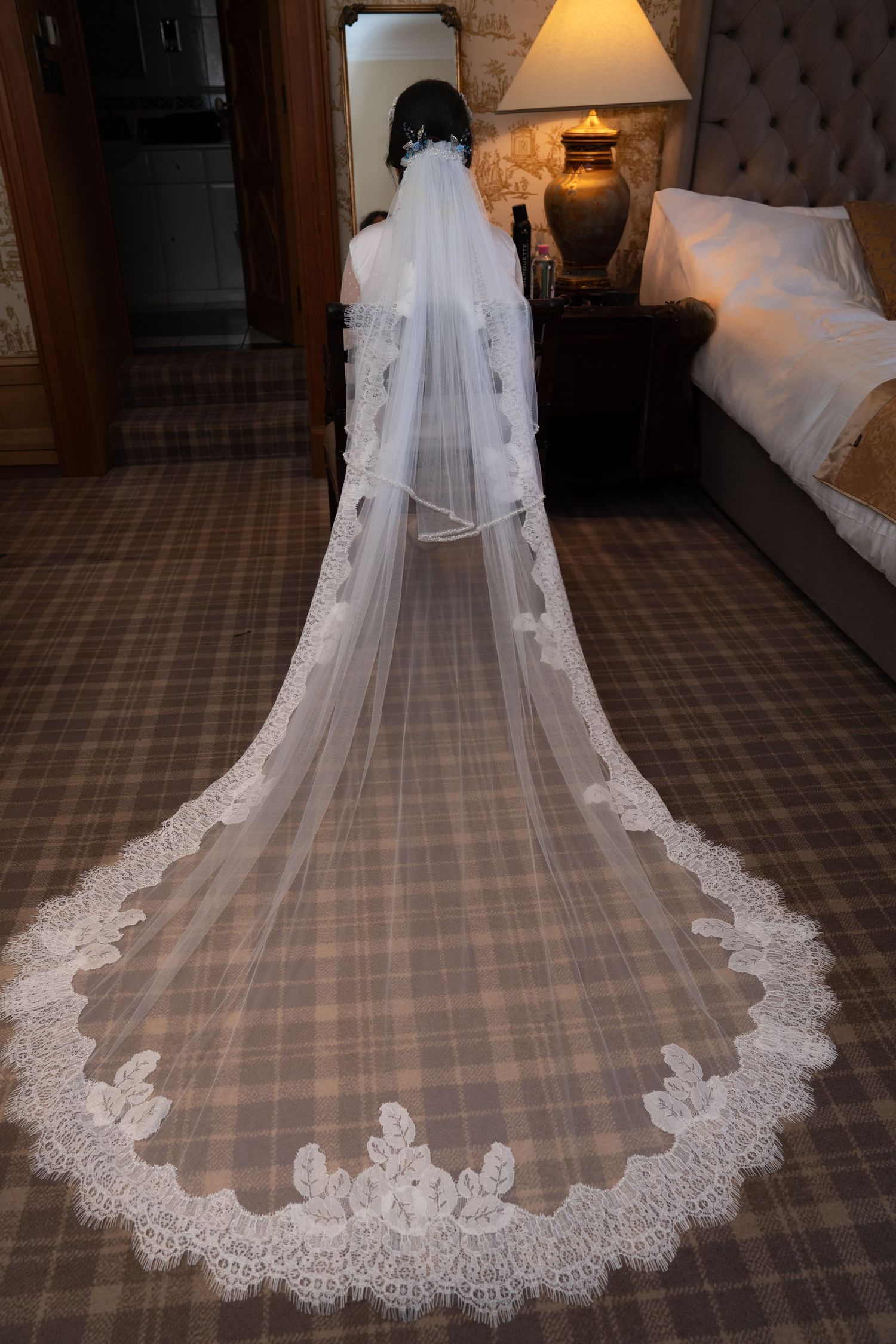 A long cathedral wedding veil with delicate lace trim lies spread out on a plaid carpeted floor next to a bedside lamp.