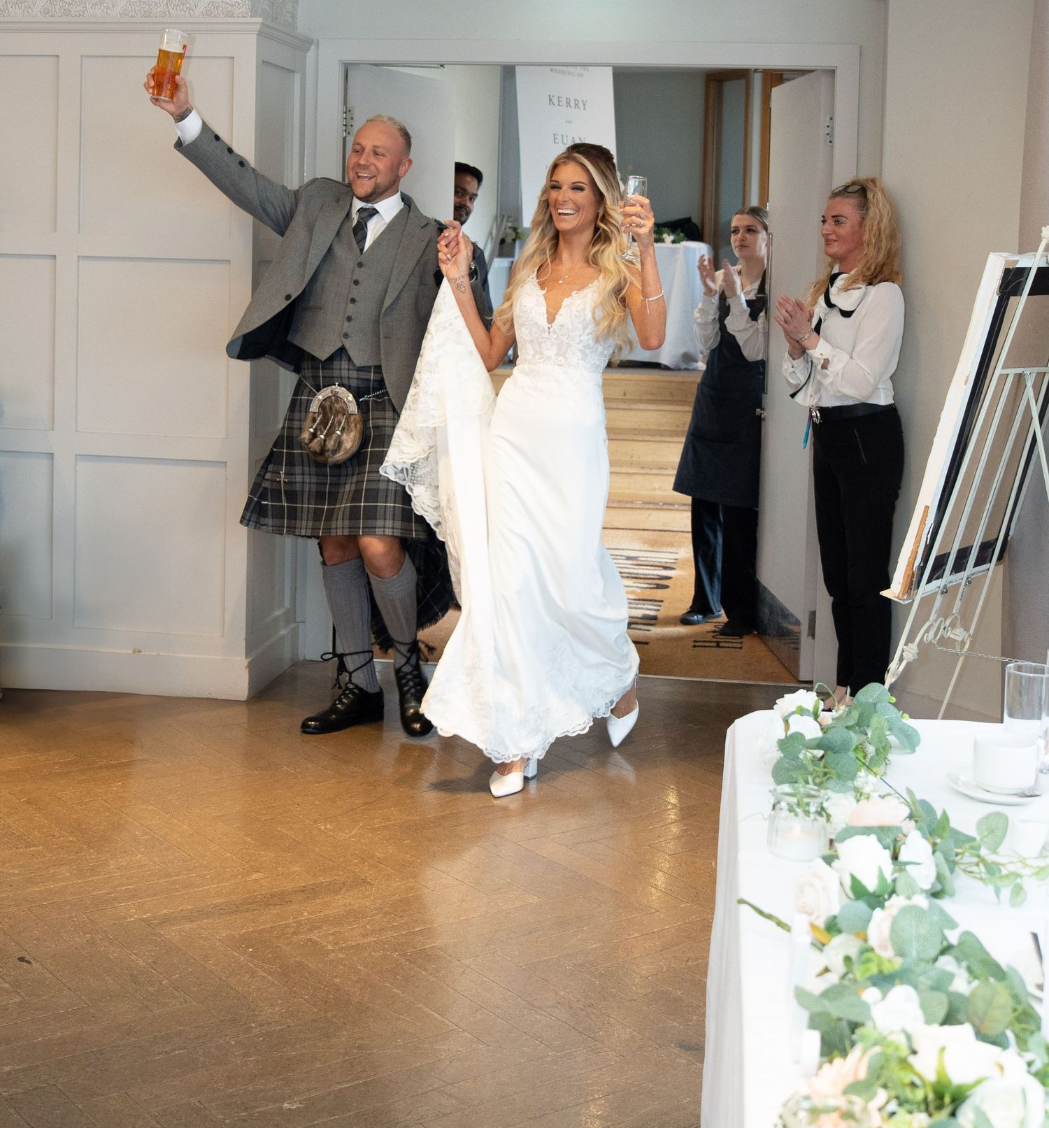 A couple celebrates at a wedding venue as they make their grand entrance, with the groom wearing a traditional kilt.