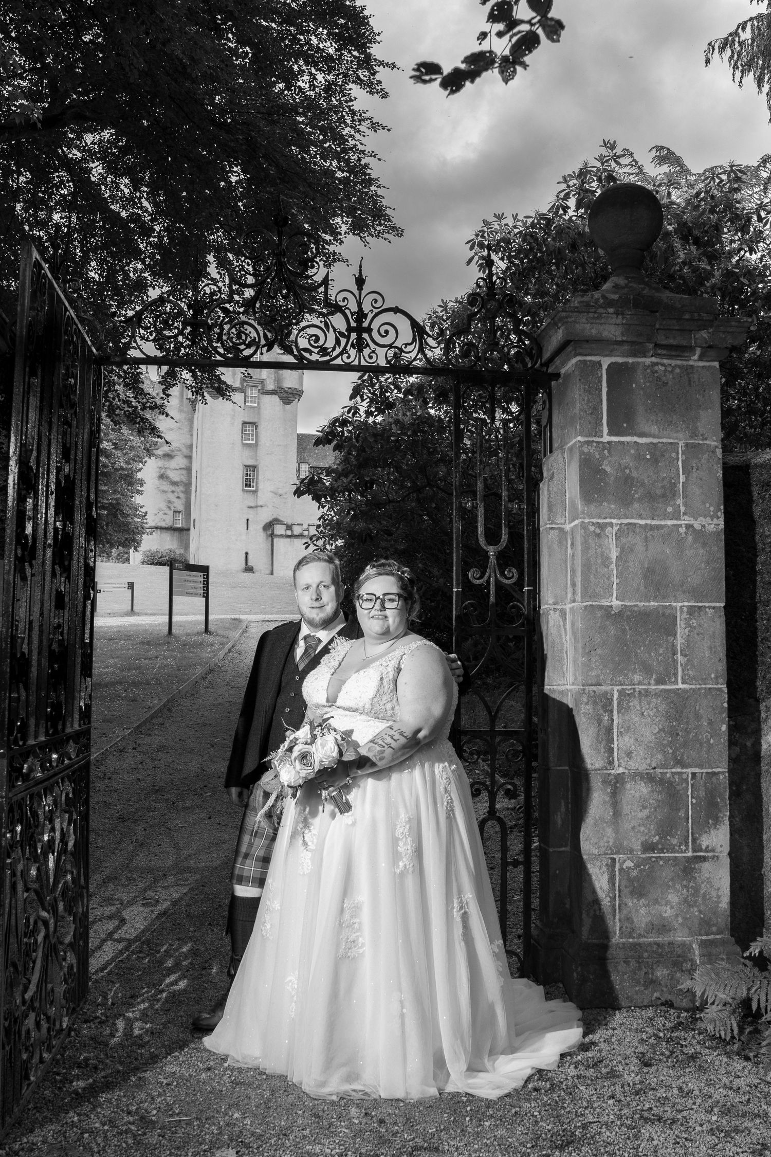 A couple poses at an ornate iron gate entrance in black and white wedding photography.