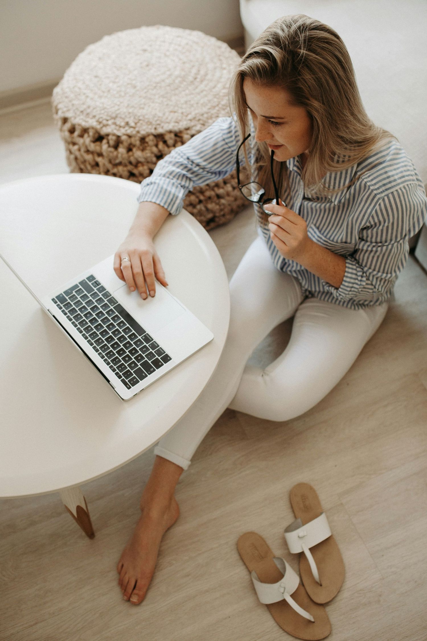 Woman in blue striped shirt and white pants working from home on laptop computer while sitting on floor.