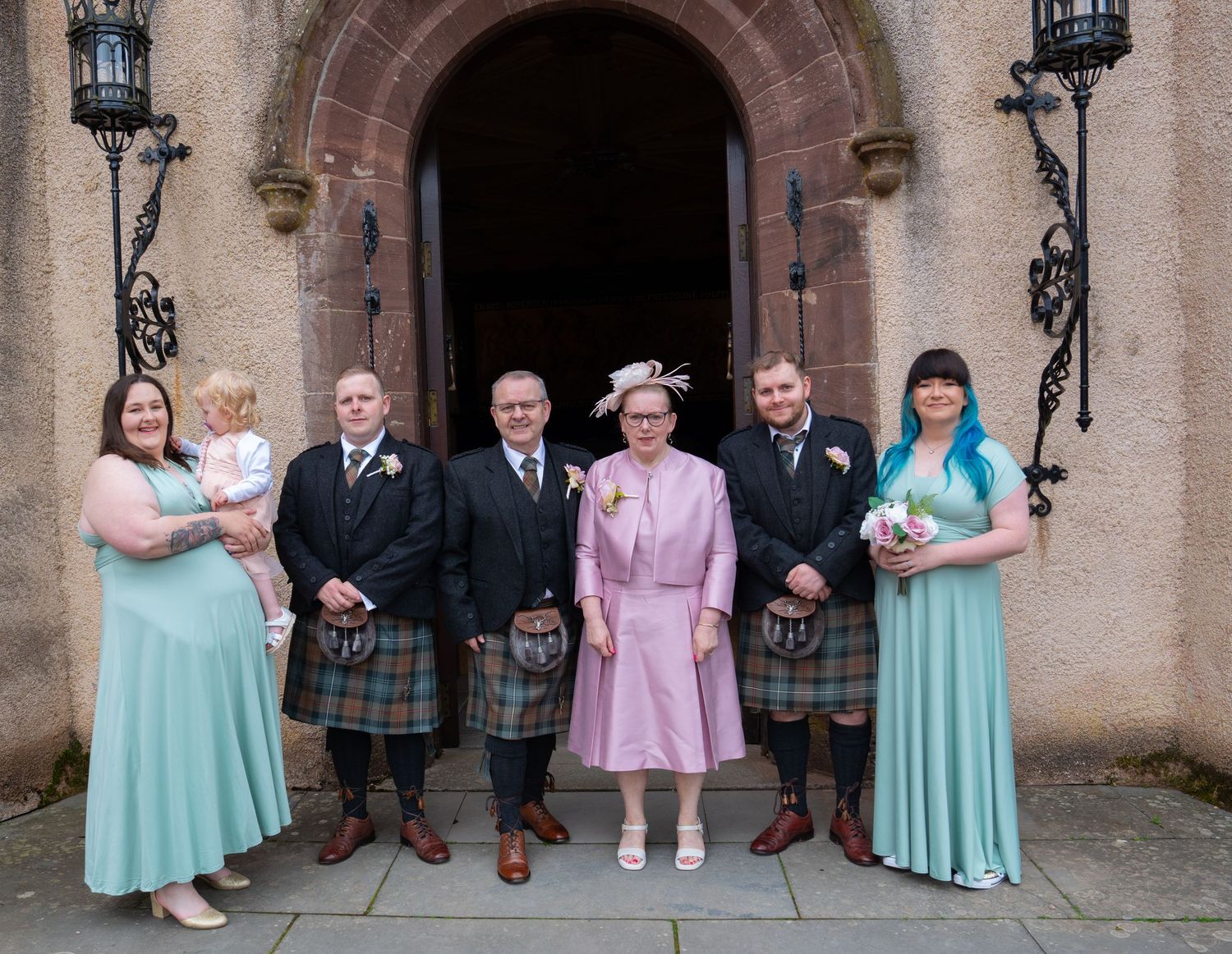 A formal group in Scottish wedding attire including kilts and long dresses pose together at a stone archway entrance.