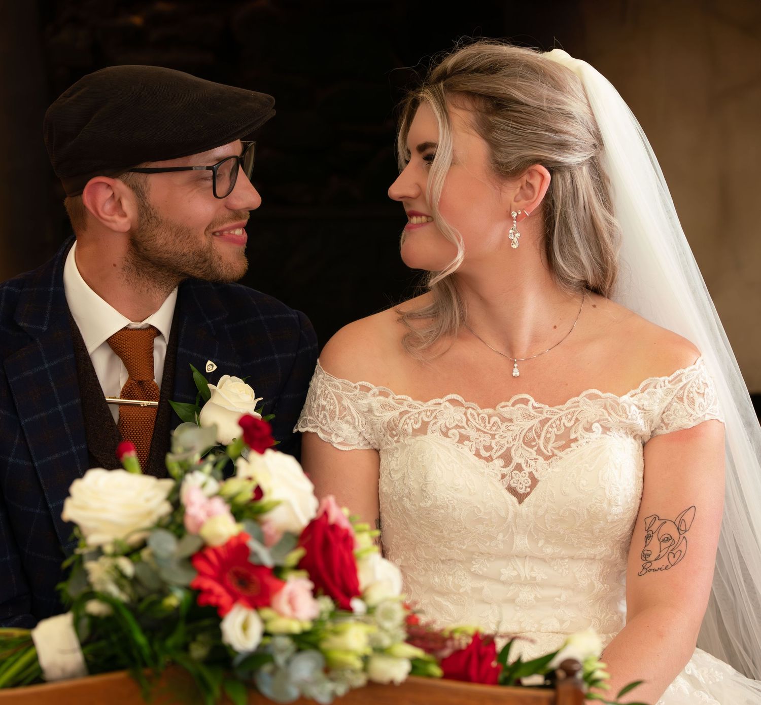 Happy couple on their wedding day smile at each other while sitting with red and white floral bouquet.